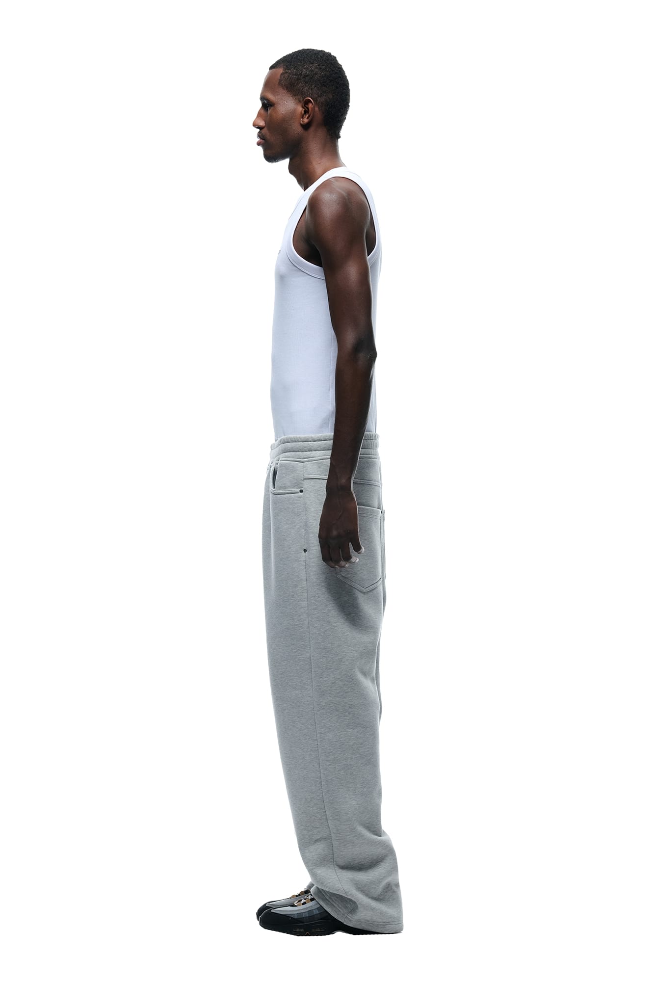 A man stands in profile against a white background, wearing a white tank top, black shoes, and 6PMs X-TRA WIDE 5-POCKET JOGGER GREY.