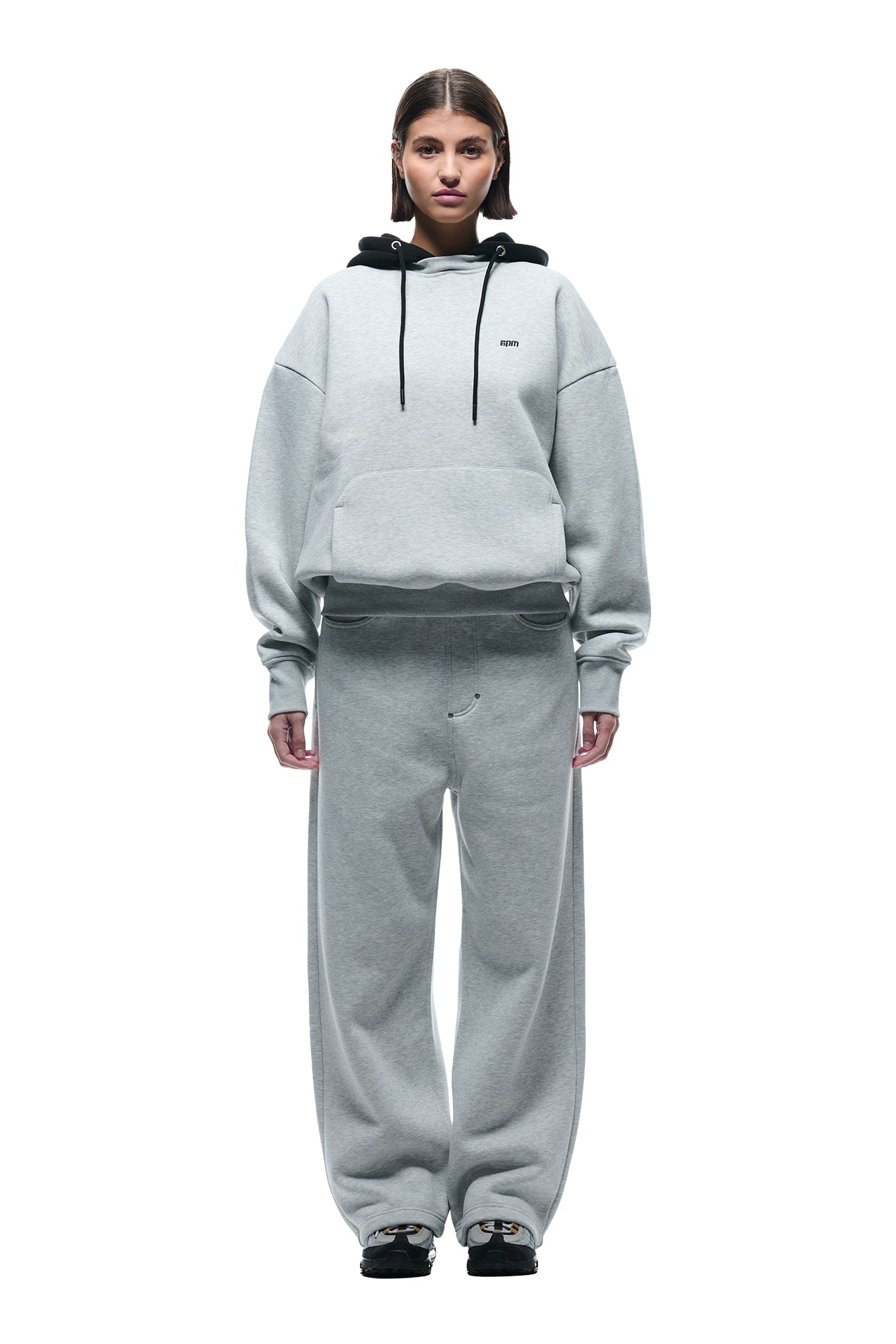 A person faces forward in a loose-fitting 6PM CREWNECK HOODIE GREY/BLACK with black drawstrings, matching wide-leg sweatpants, and black sneakers. The background is plain white.