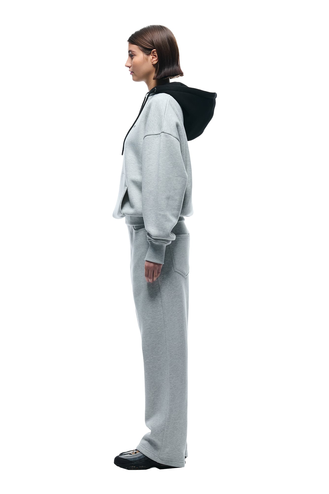 A woman with straight brown hair stands in profile against a white background, wearing the 6PM CREWNECK HOODIE GREY/BLACK and matching sweatpants with black shoes.