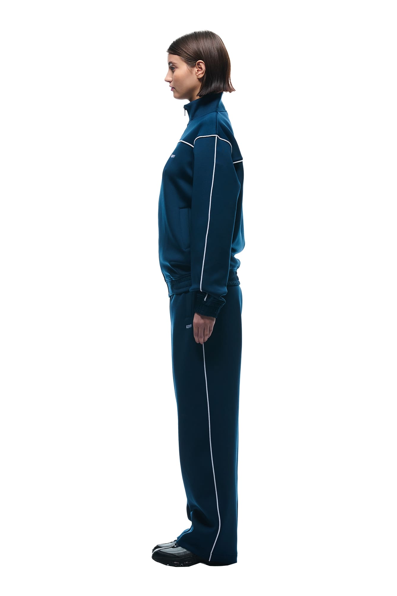 A person with short brown hair stands in profile against a plain white background, wearing the 6PM NEOPRENE TRACK JACKET NAVY and black shoes.