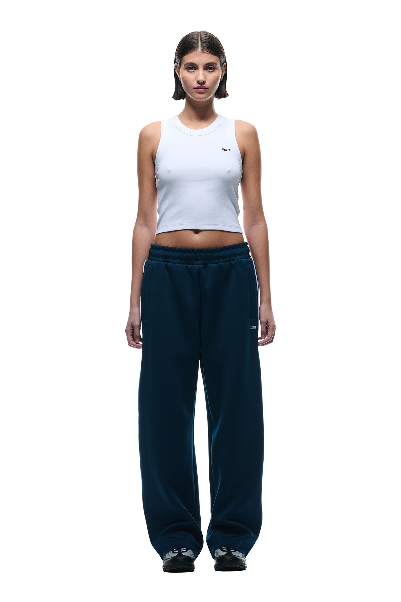 A woman faces forward against a plain white background, wearing 6PMs NEOPRENE TRACK PANTS in navy with a white sleeveless crop top and black sneakers.