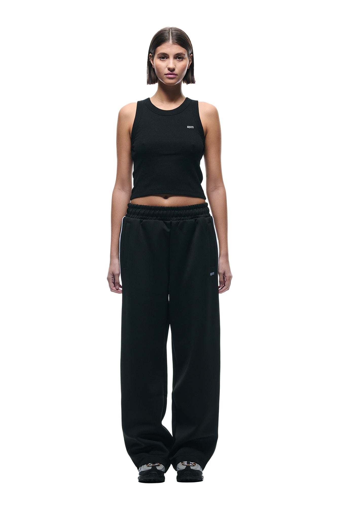 A woman with straight, shoulder-length hair wears 6PMs NEOPRENE TRACK PANTS BLACK, a black sleeveless crop top, and black sneakers, standing against a plain white background.