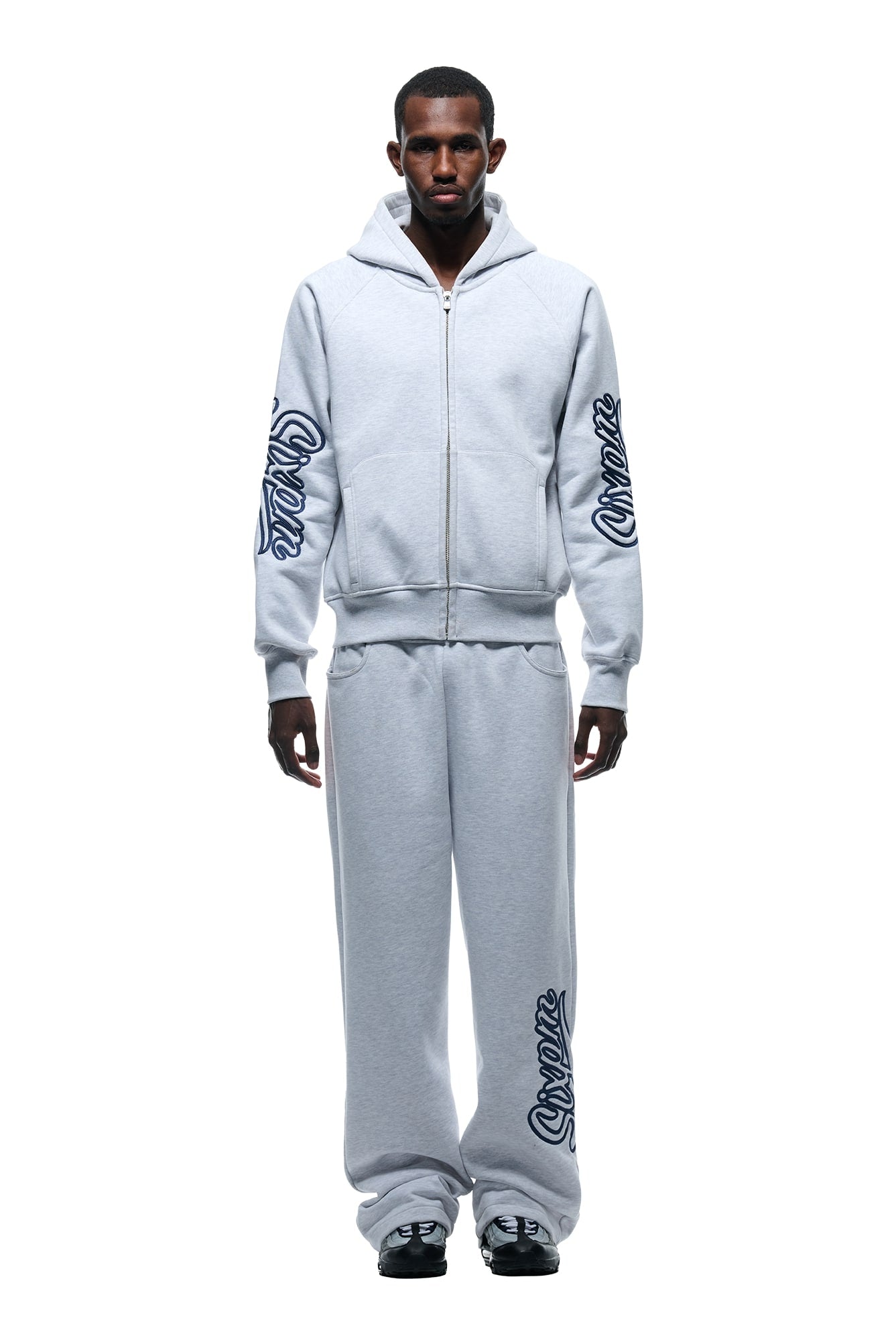 A man faces forward in a light grey WAVE LOGO ZIP HOODIE from 6PM, featuring bold blue script on the sleeves, paired with matching sweatpants and black-and-white sneakers against a plain white background.