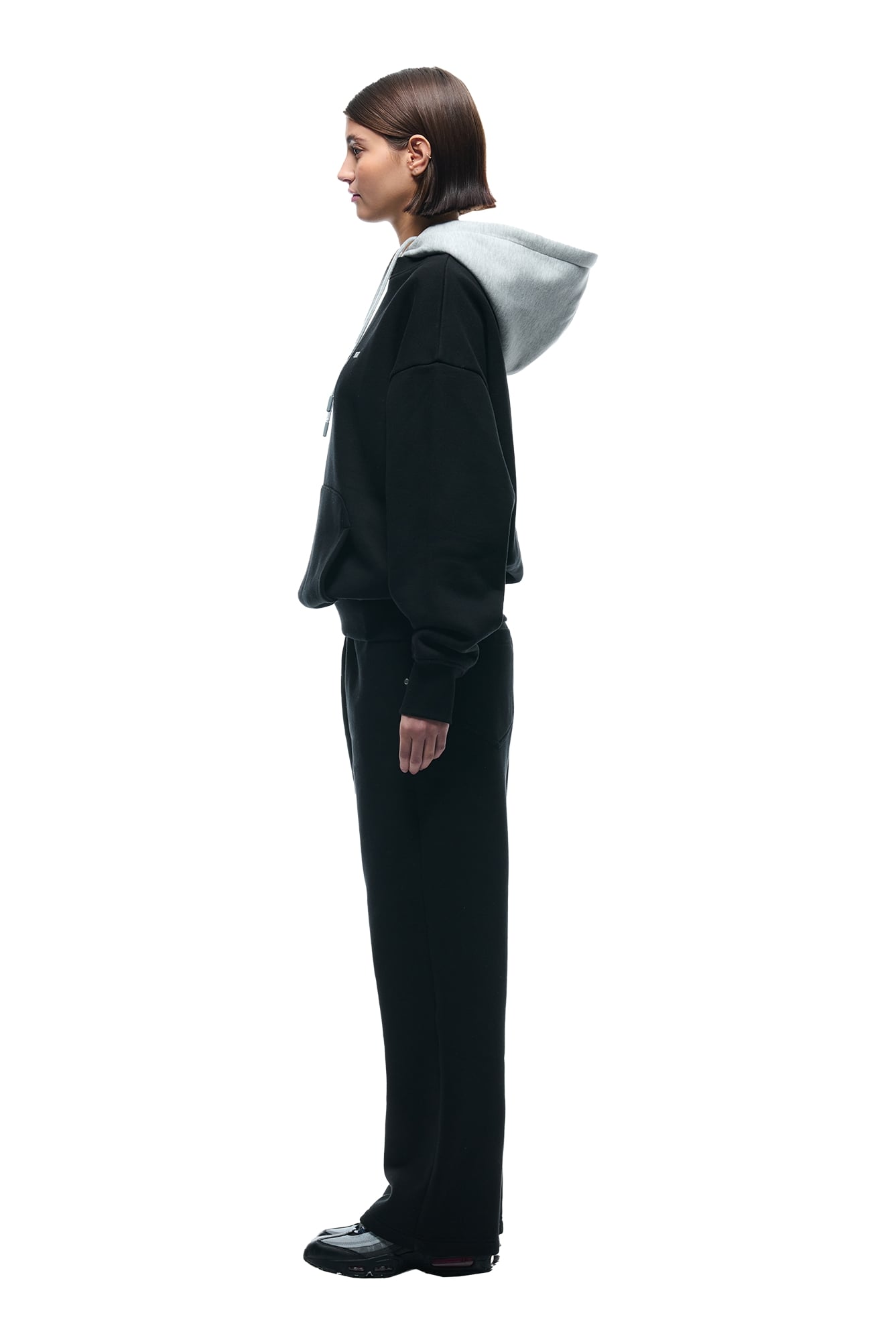 A person with straight brown hair stands in profile against a white background, wearing the 6PM CREWNECK HOODIE BLACK/GREY, paired with black pants and black shoes.