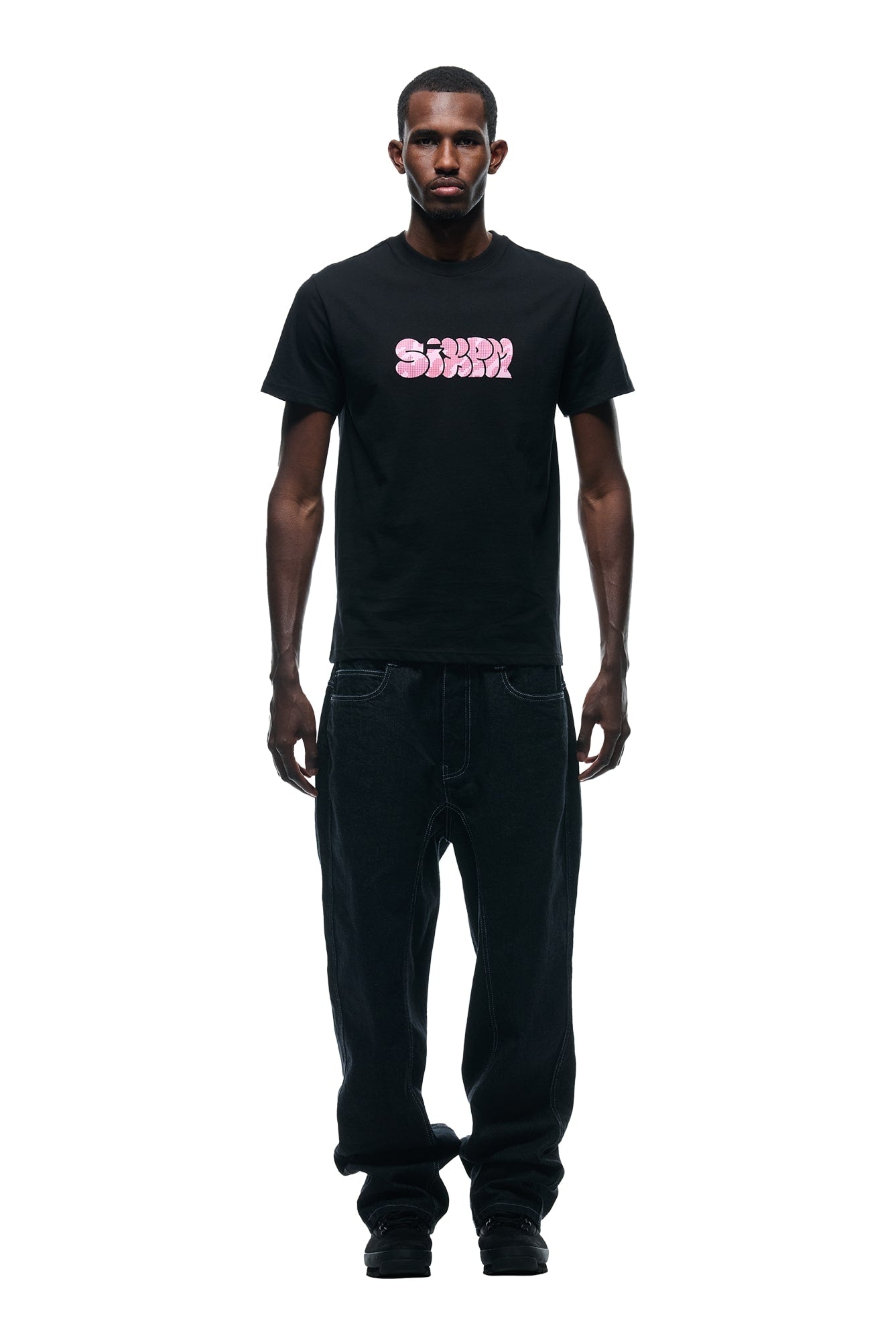 A man faces forward against a white background, wearing the 6PM GRAFITTY CAMO T-SHIRT BLACK with bold pink Sorry Sorry text, paired with loose black jeans and black shoes.