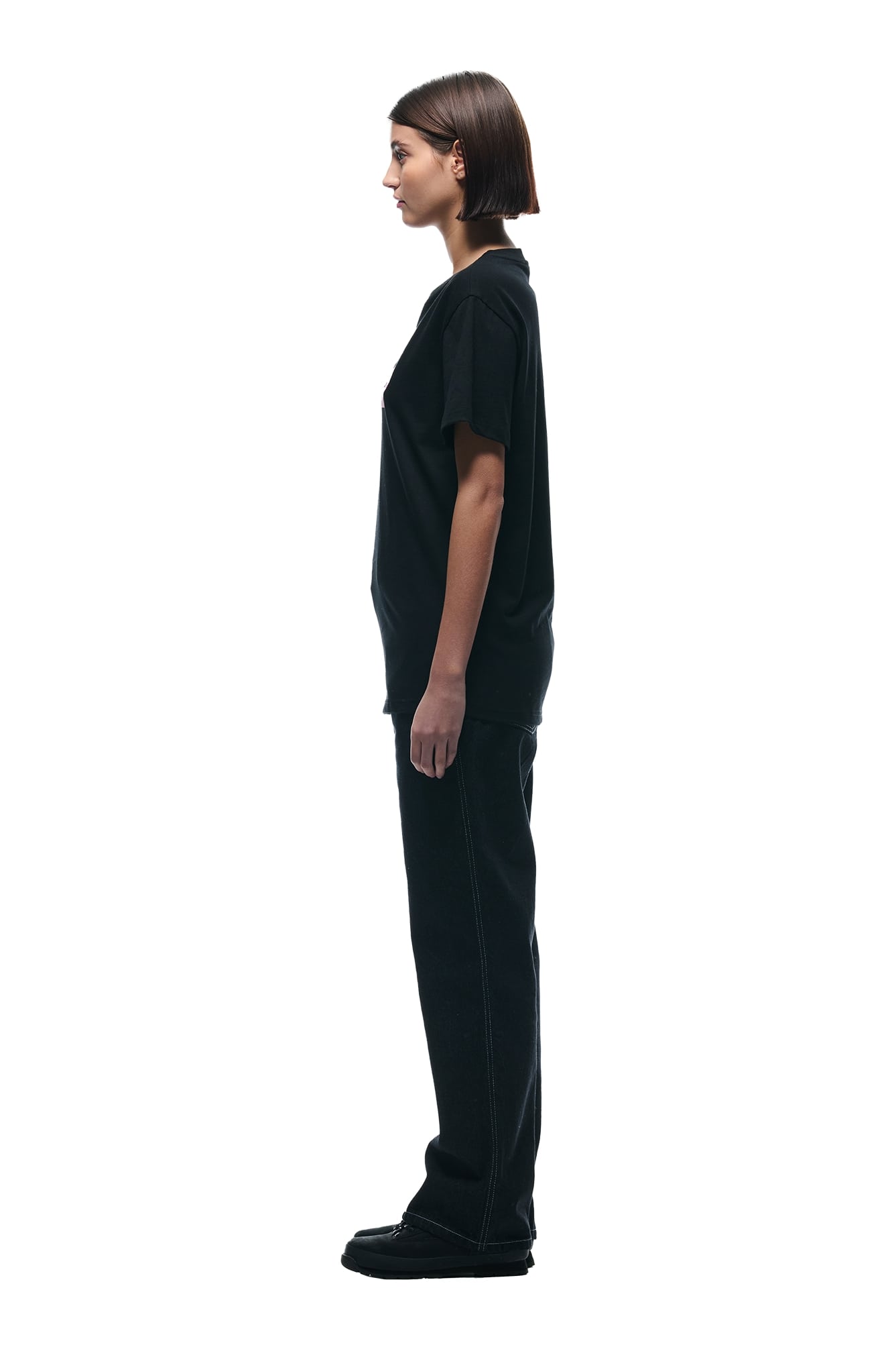 A person with short brown hair stands in profile facing left, wearing the 6PM GRAFITTY CAMO T-SHIRT BLACK, black pants, and black shoes against a plain white background.