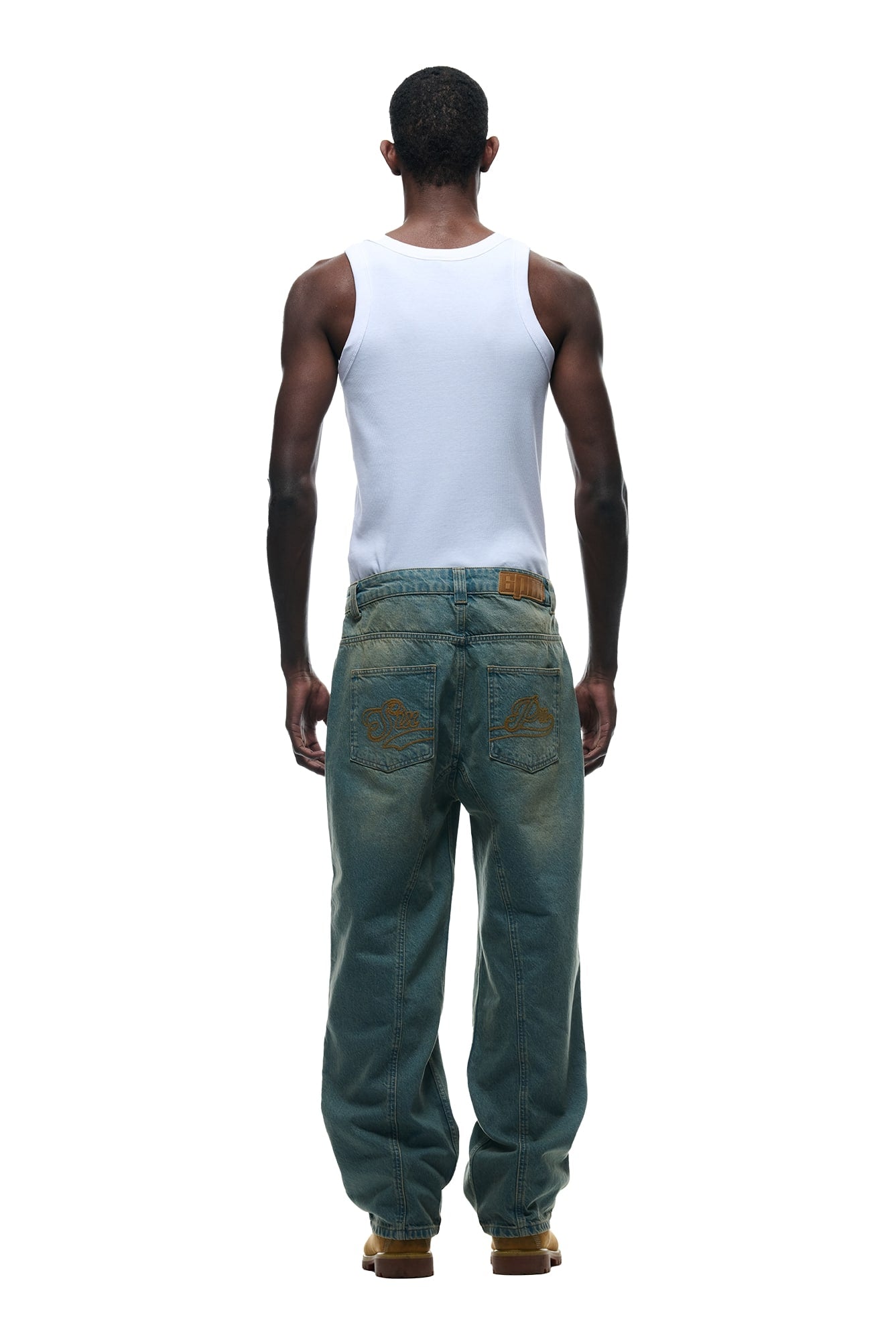 A man faces away against a plain white background, wearing a white tank top, tan boots, and 6PM CENTER SEAM DENIM LIGHT WASH jeans with designed back pockets.
