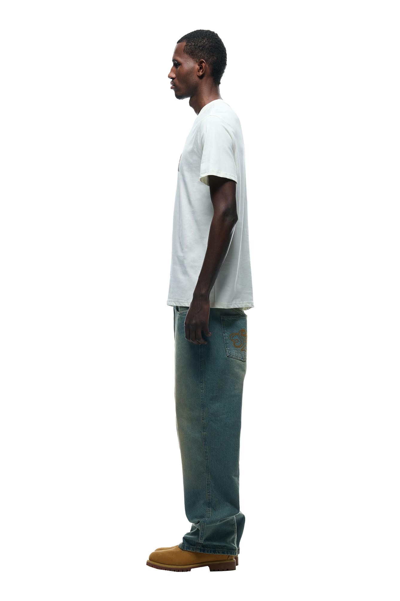 A man stands in profile against a white background, wearing the 6PM GRAFITTY CAMO T-SHIRT MARSHMALLOW, blue jeans with a graphic on the pocket, and tan boots.