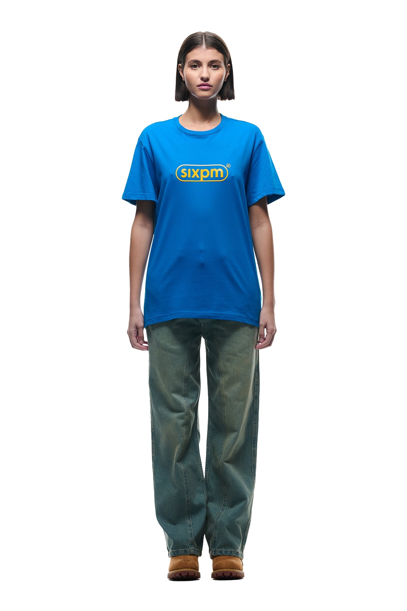 A person with short brown hair wears a 6PM DURSIX TEE in Princess Blue, loose green pants, and brown shoes, standing facing forward against a plain white background.