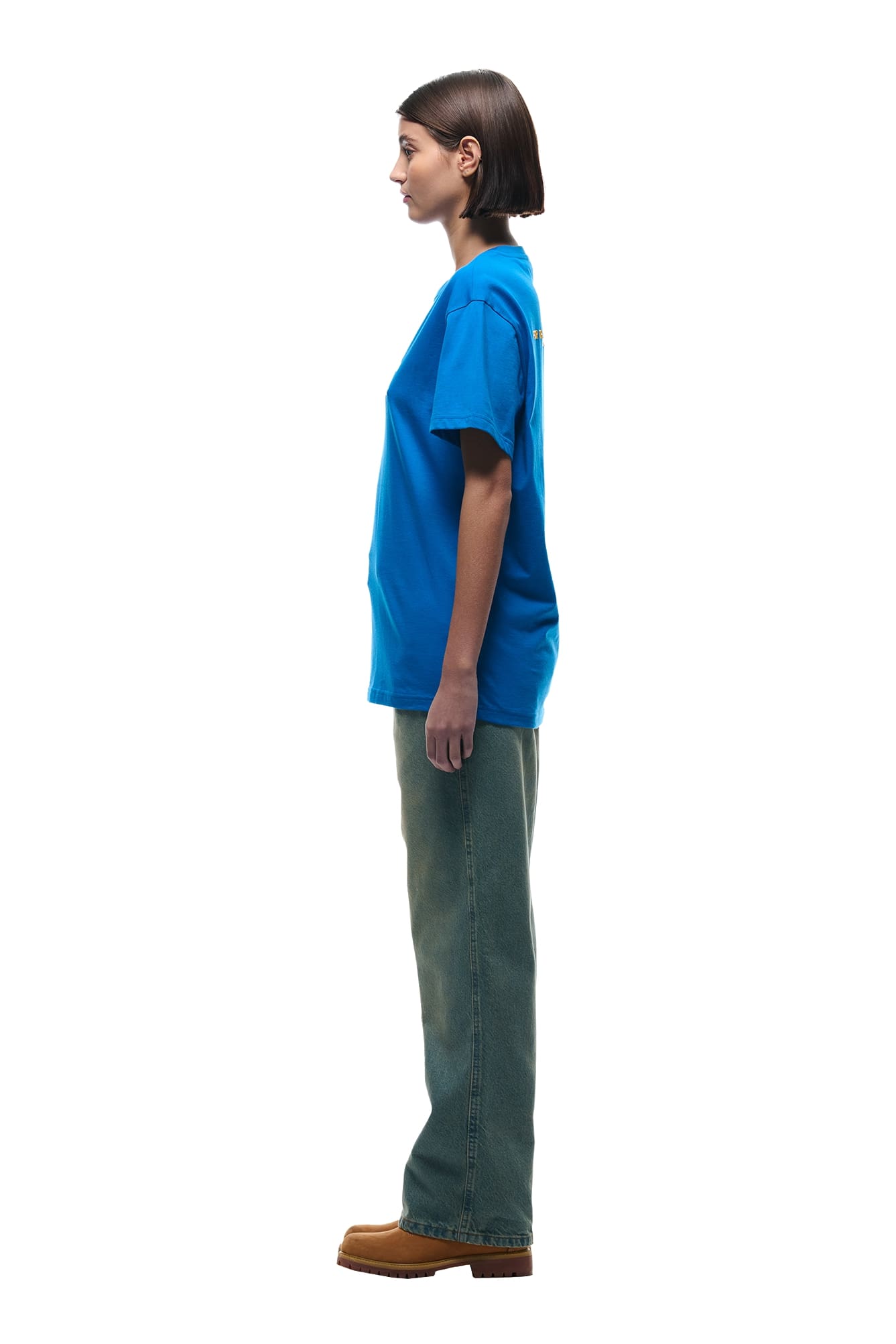 A person with short brown hair stands in profile facing left, wearing the 6PM DURSIX TEE PRINCESS BLUE, greenish jeans, and brown shoes against a white background.