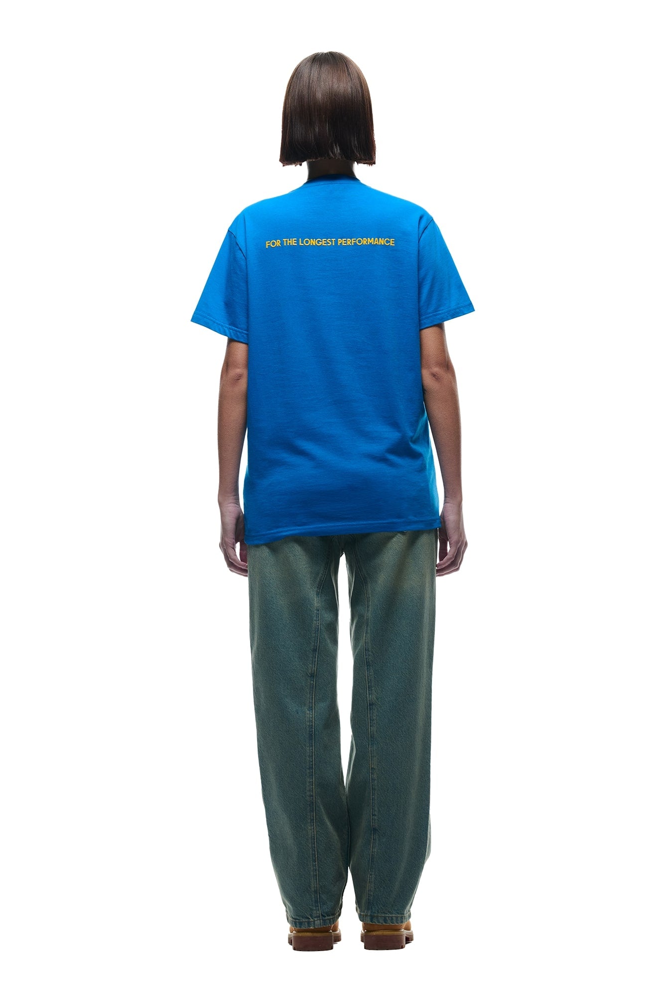 A person with short hair faces away, wearing the 6PM DURSIX TEE PRINCESS BLUE featuring yellow FOR THE LONGEST PERFORMANCE text on the back, loose blue jeans, and brown shoes, set against a white background.