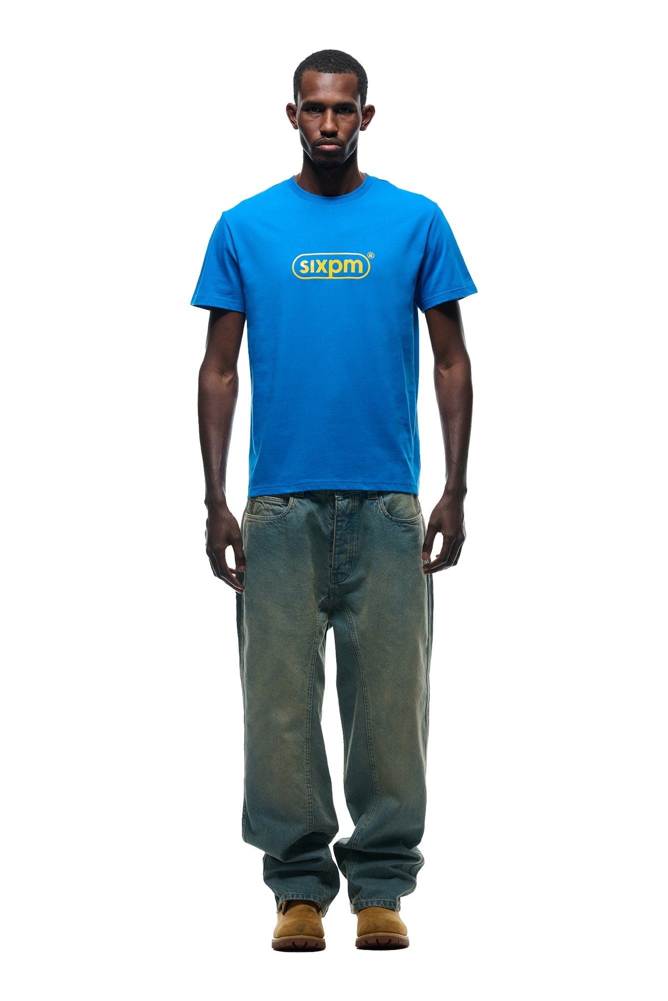 A man faces forward against a white background, wearing the 6PM DURSIX TEE PRINCESS BLUE, loose-fitting faded jeans, and tan shoes.