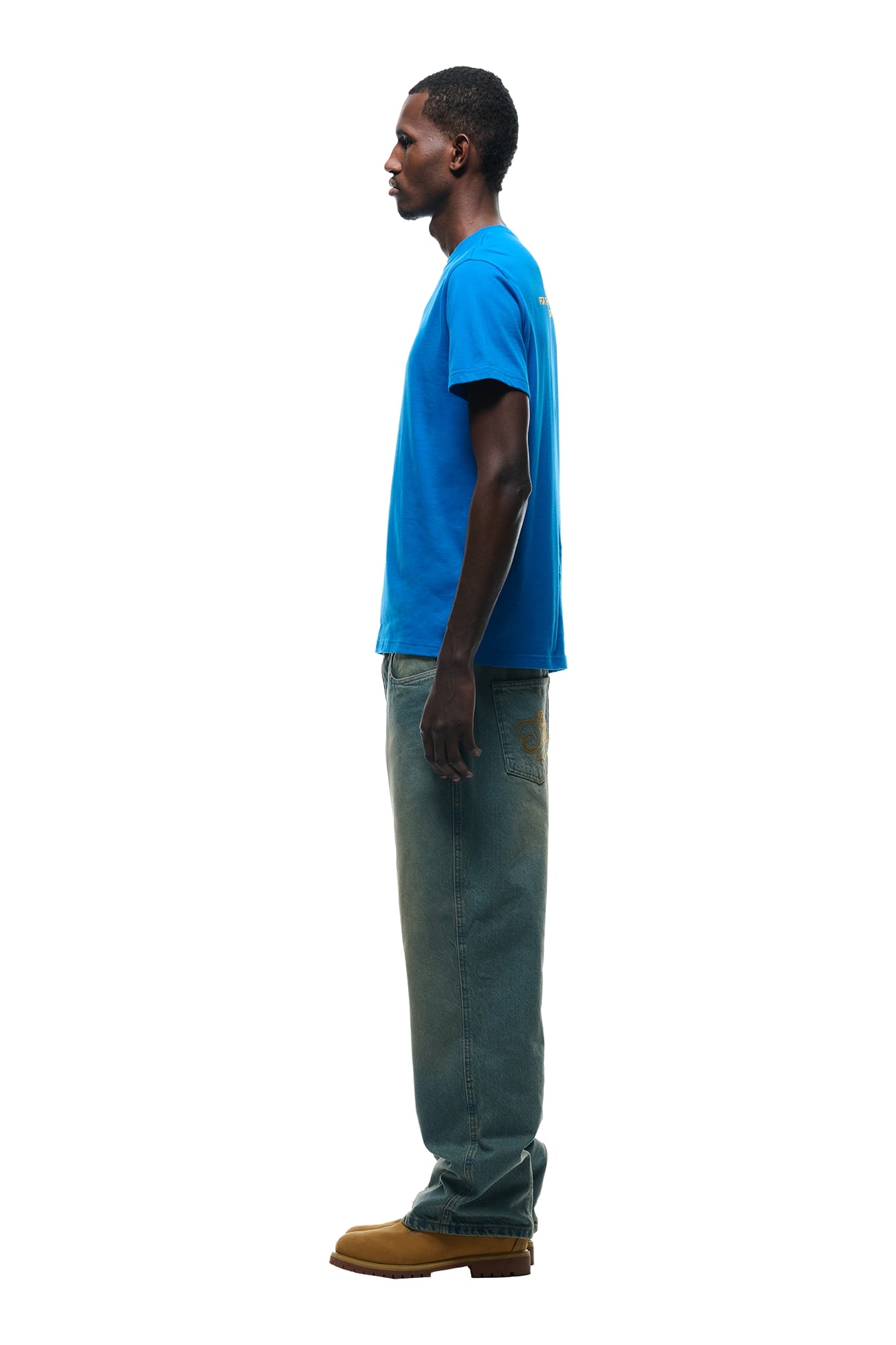 A man stands in profile against a white background, wearing the 6PM DURSIX TEE PRINCESS BLUE, loose-fitting blue jeans, and tan work boots.