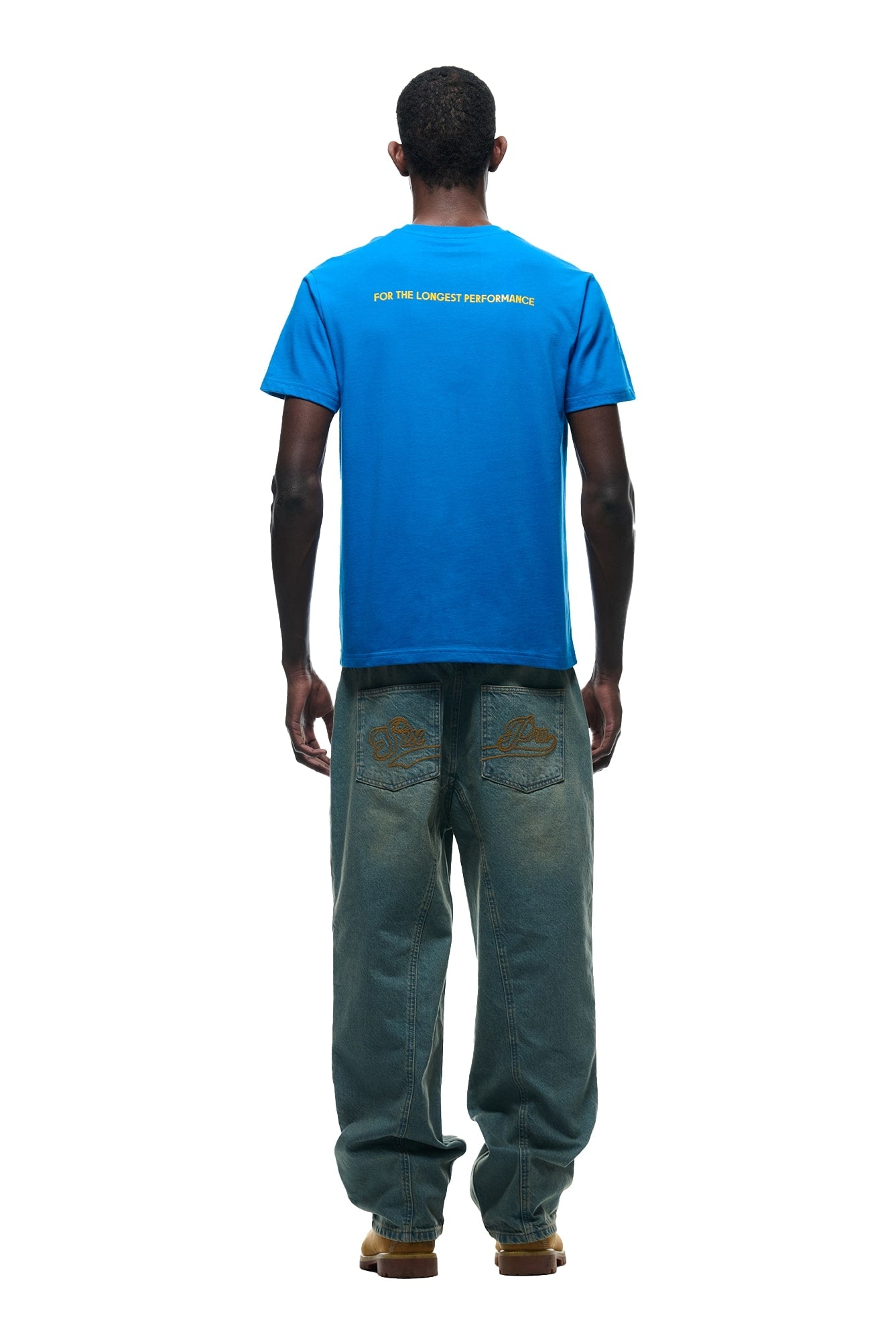 A person stands with their back to the camera, wearing the 6PM DURSIX TEE PRINCESS BLUE with white FOR THE LONGEST PERFORMANCE text, loose blue jeans featuring back pocket designs, and tan shoes, set against a white background.