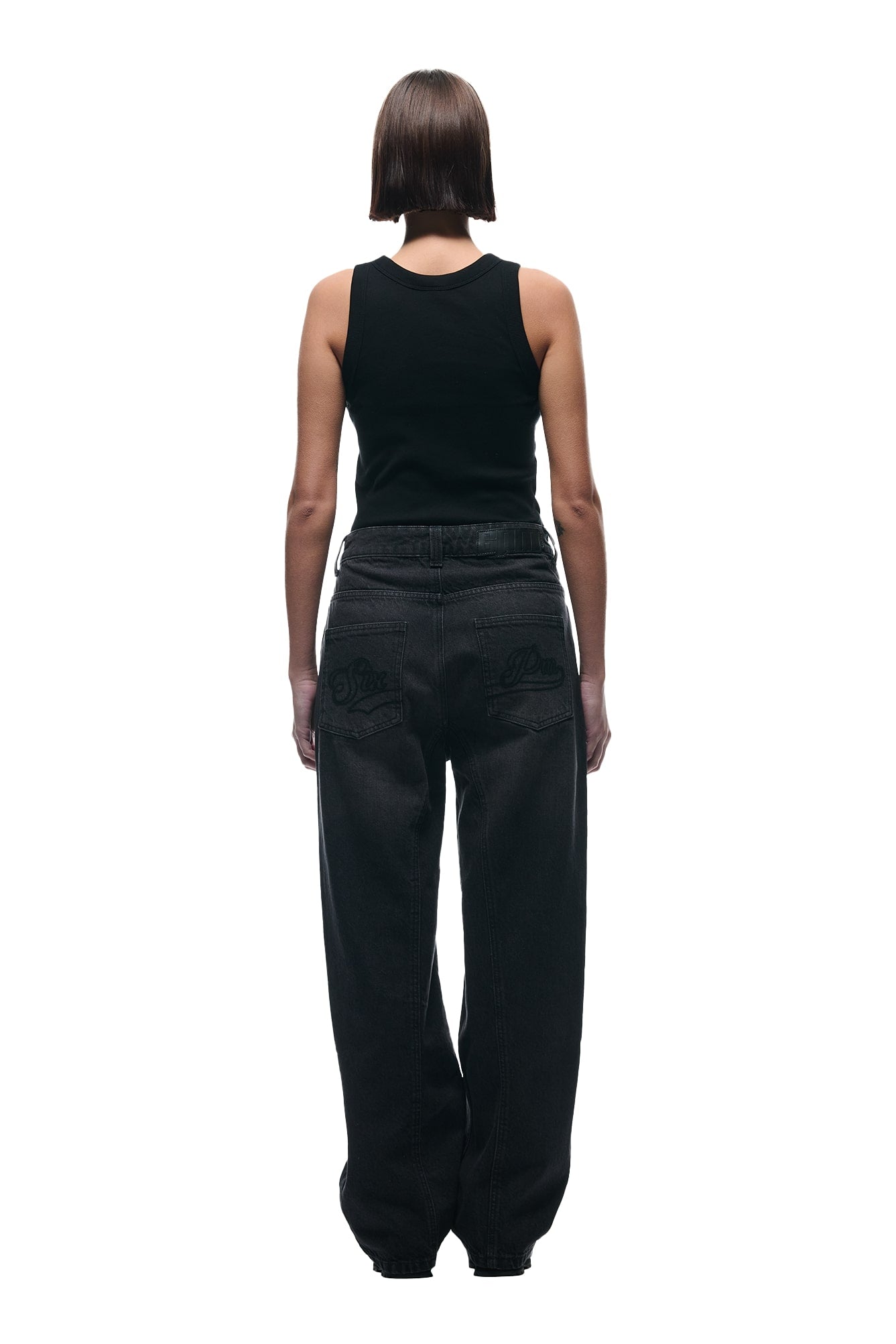 A person with straight, chin-length hair faces away from the camera, wearing a black sleeveless top and 6PM’s CENTER SEAM DENIM PURPLE OVERDYE jeans with decorative back pocket stitching, set against a white background.
