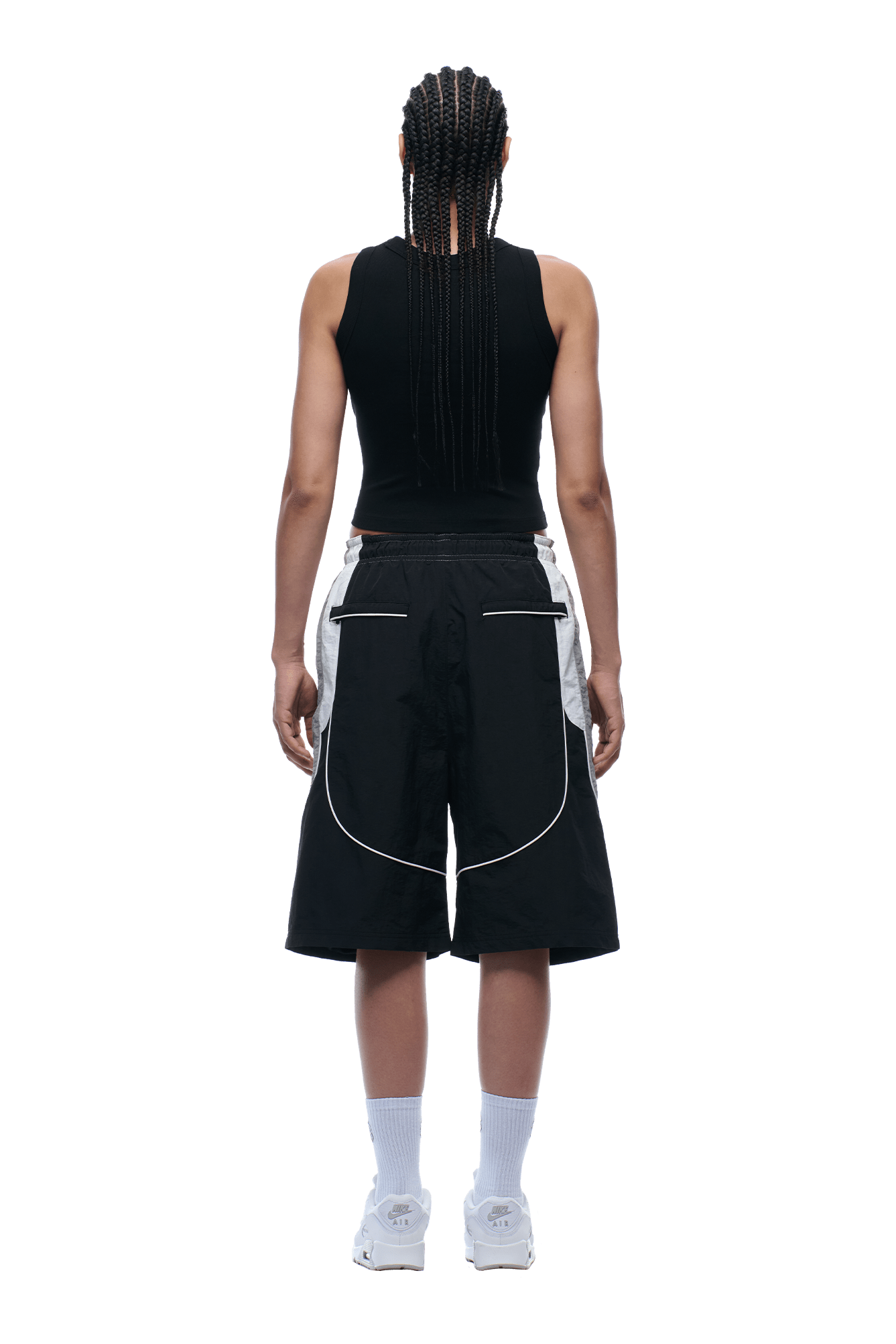 A person with long braids stands facing away from the camera, wearing a black sleeveless top, 6PM BAGGY WARM UP SHORTS BLACK, white-trimmed black shorts, white socks, and white sneakers against a transparent background.