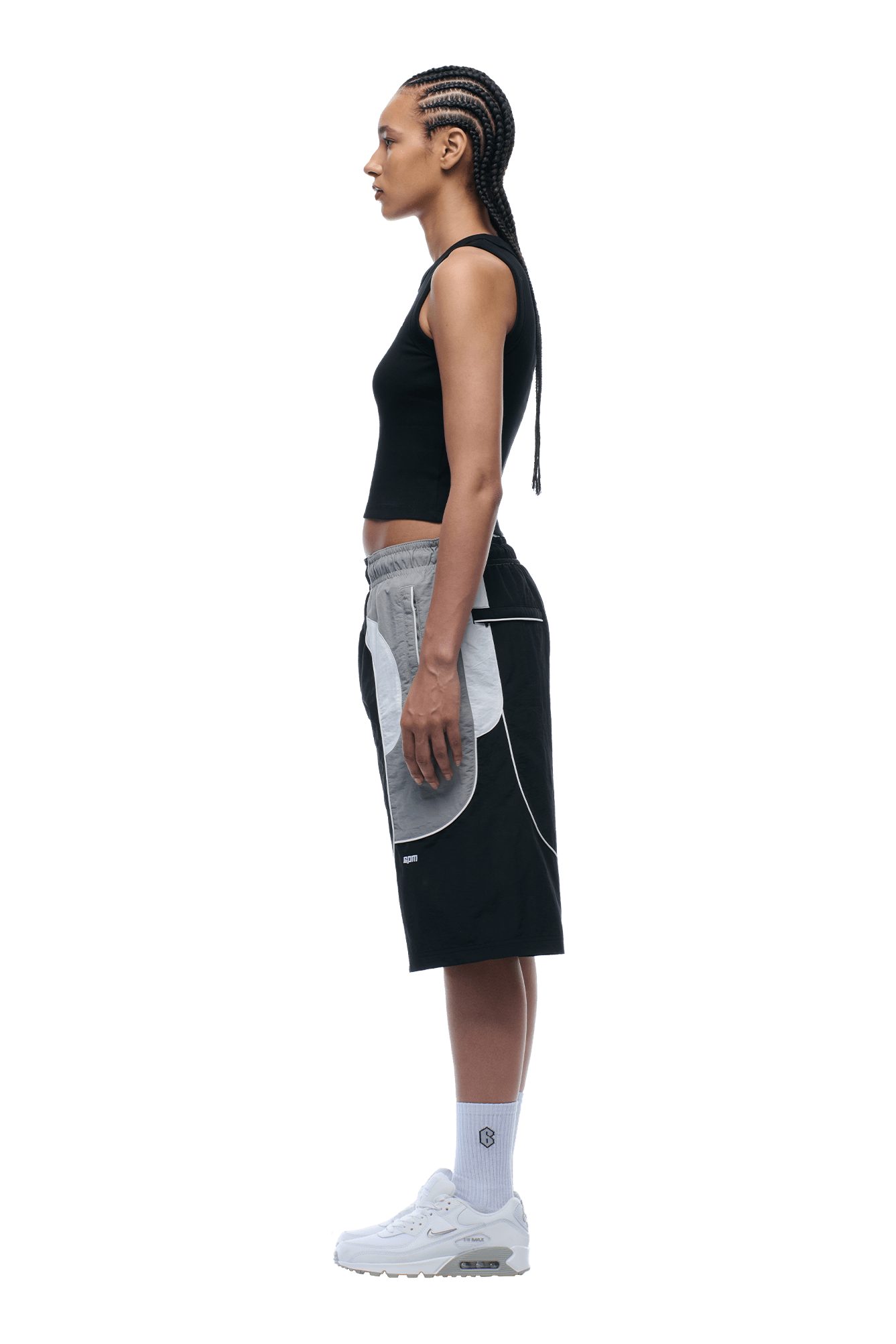 A woman with braided hair stands in profile against a plain background, wearing a black sleeveless top, 6PM BAGGY WARM UP SHORTS BLACK with gray and white panels, plus white crew socks and sneakers.