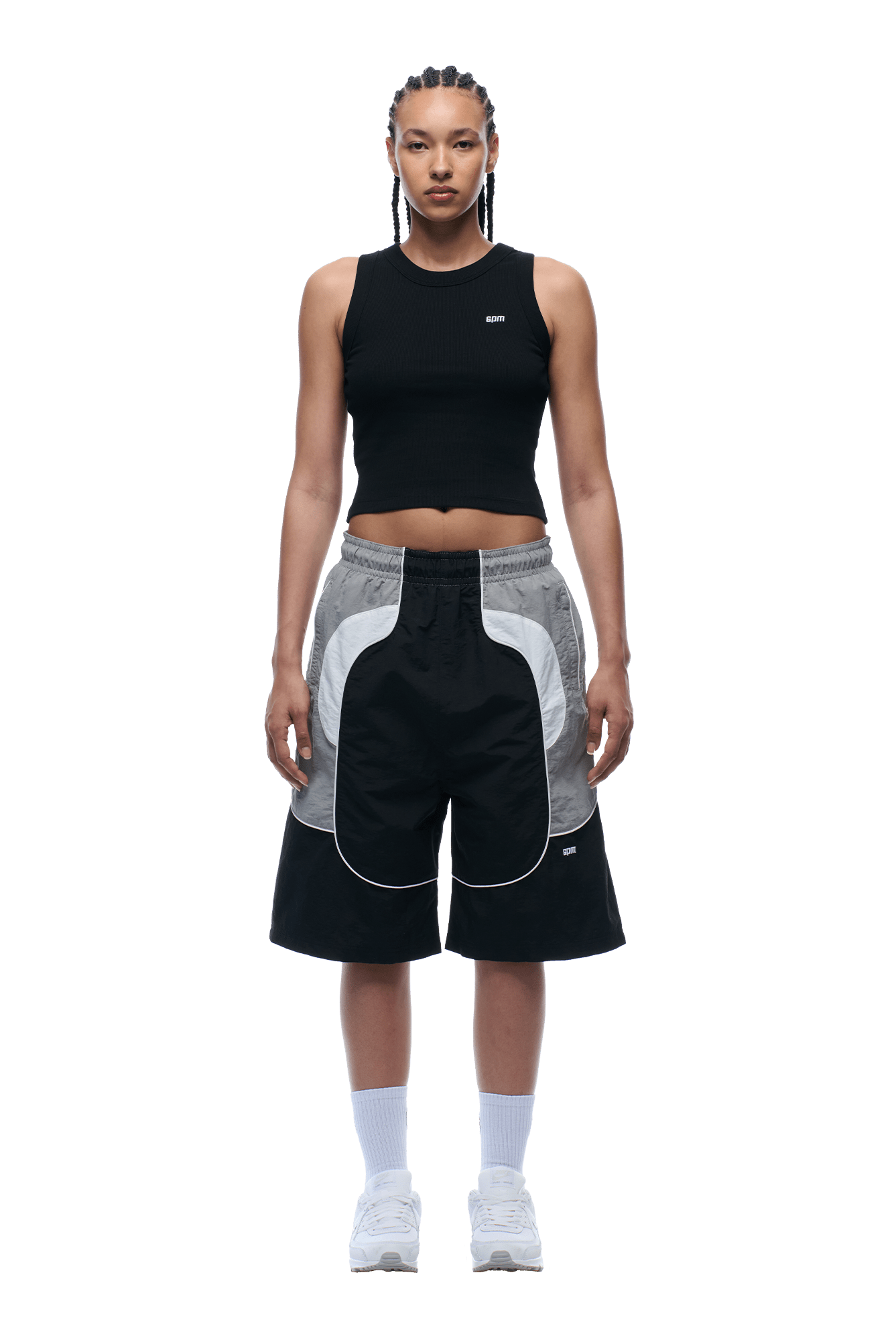 A person with braided hair wears a black sleeveless crop top, 6PM BAGGY WARM UP SHORTS BLACK, white socks, and white sneakers, standing facing forward against a plain background.