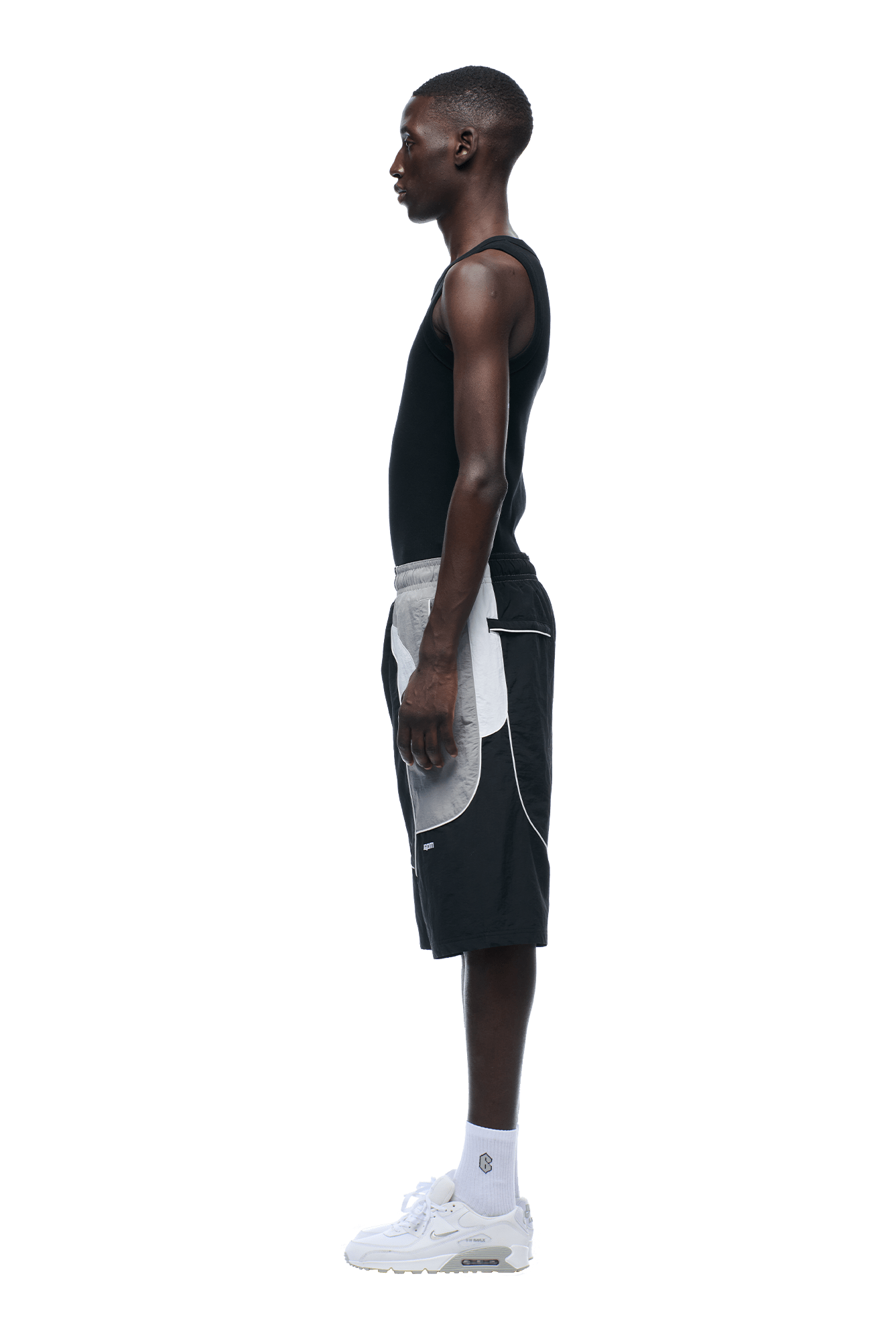 A young man stands in profile facing left, wearing a black tank top, 6PM BAGGY WARM UP SHORTS BLACK, white socks, and white sneakers against a plain background.
