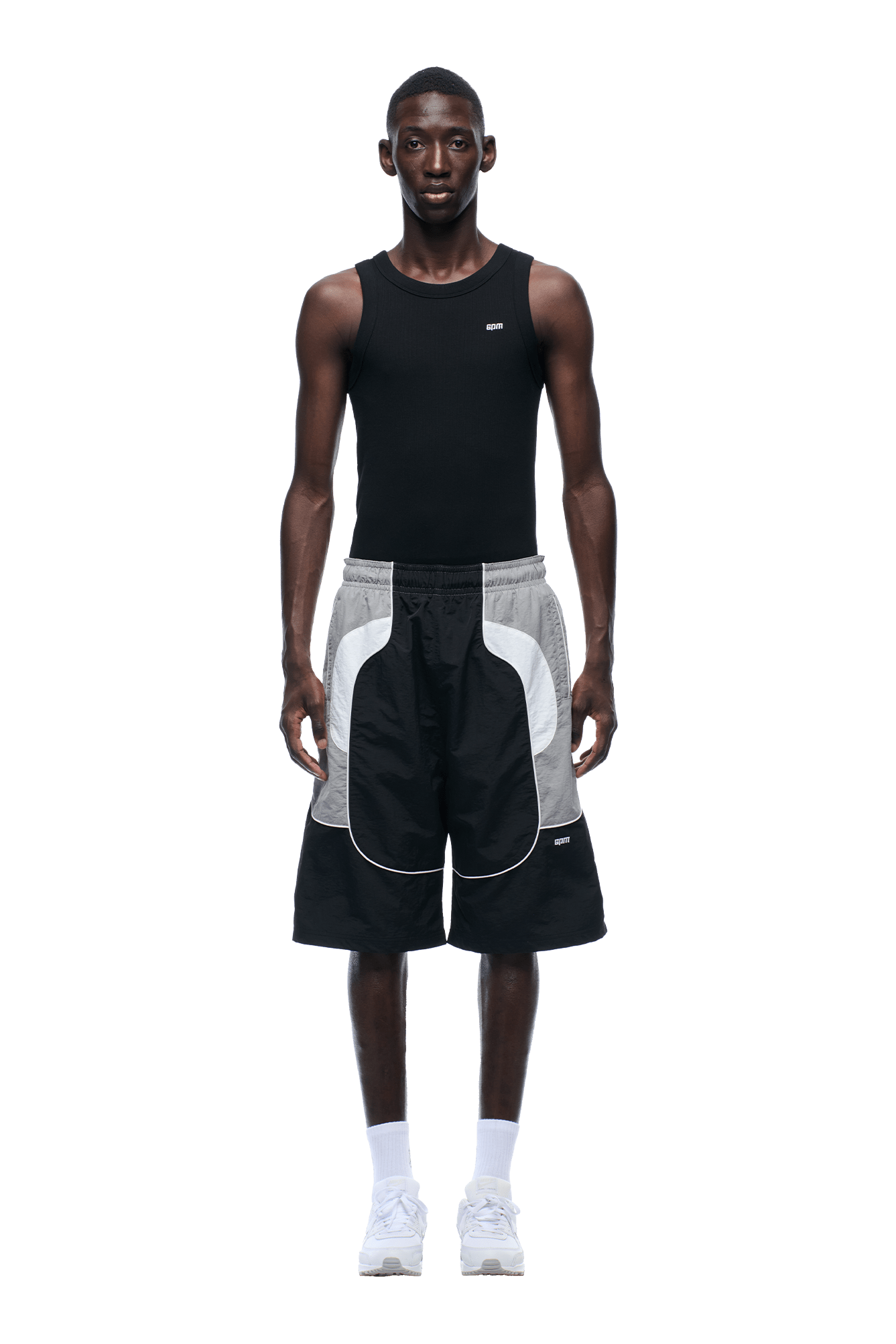A man stands facing forward, wearing a sleeveless black shirt, 6PM BAGGY WARM UP SHORTS BLACK, white socks, and white sneakers against a plain background.