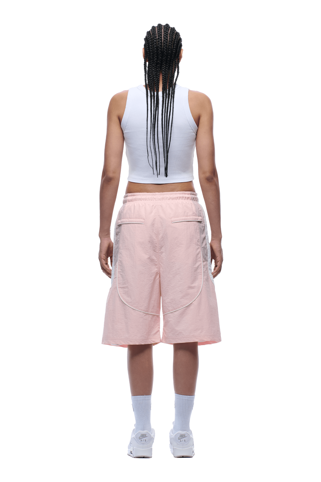 A person with long braids faces away, wearing a white sleeveless crop top, 6PM BAGGY WARM UP SHORTS PINK, white socks, and white sneakers against a plain background.