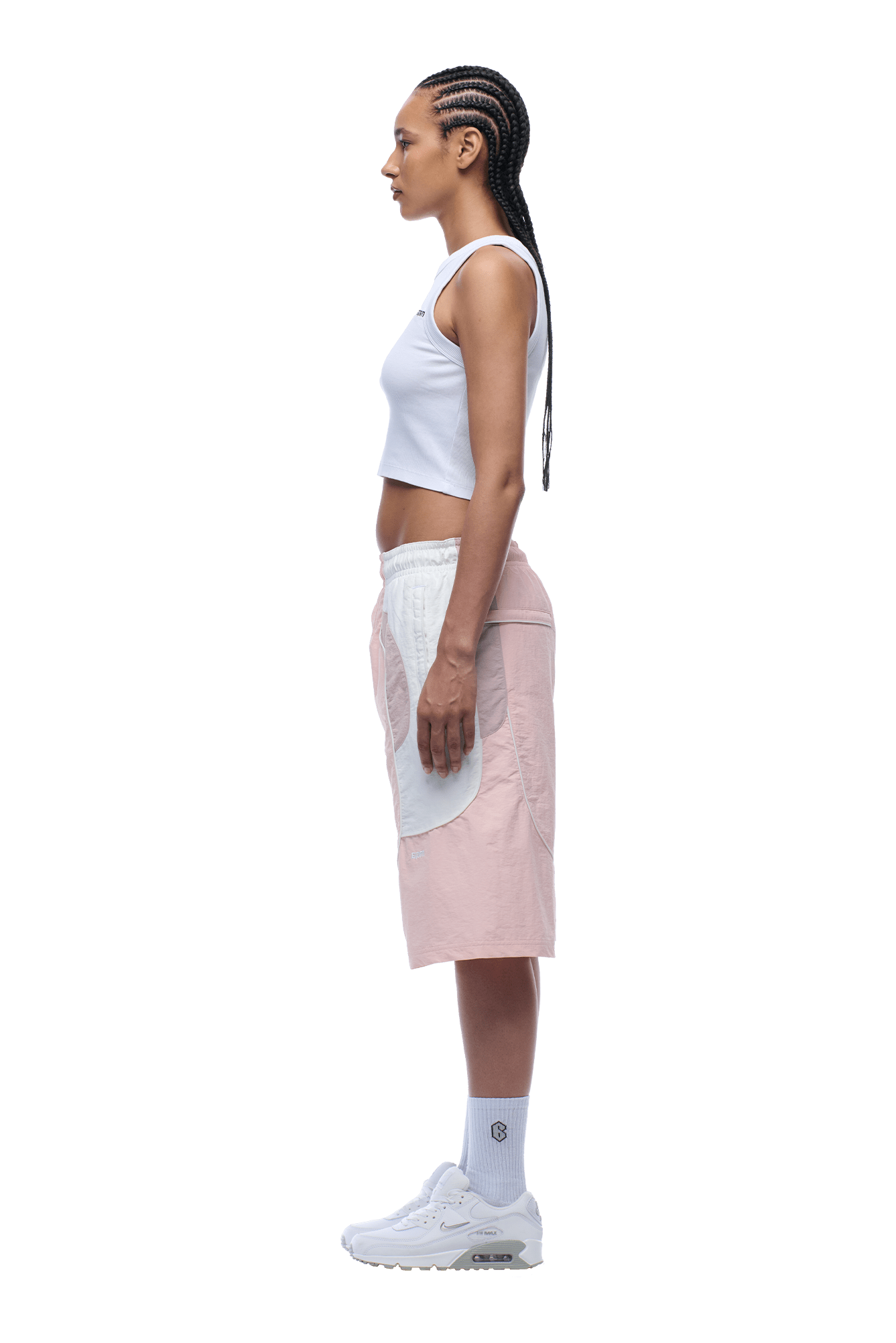 A person with braided hair stands in profile against a plain background, wearing a white crop top, 6PM BAGGY WARM UP SHORTS PINK, white socks, and white sneakers.