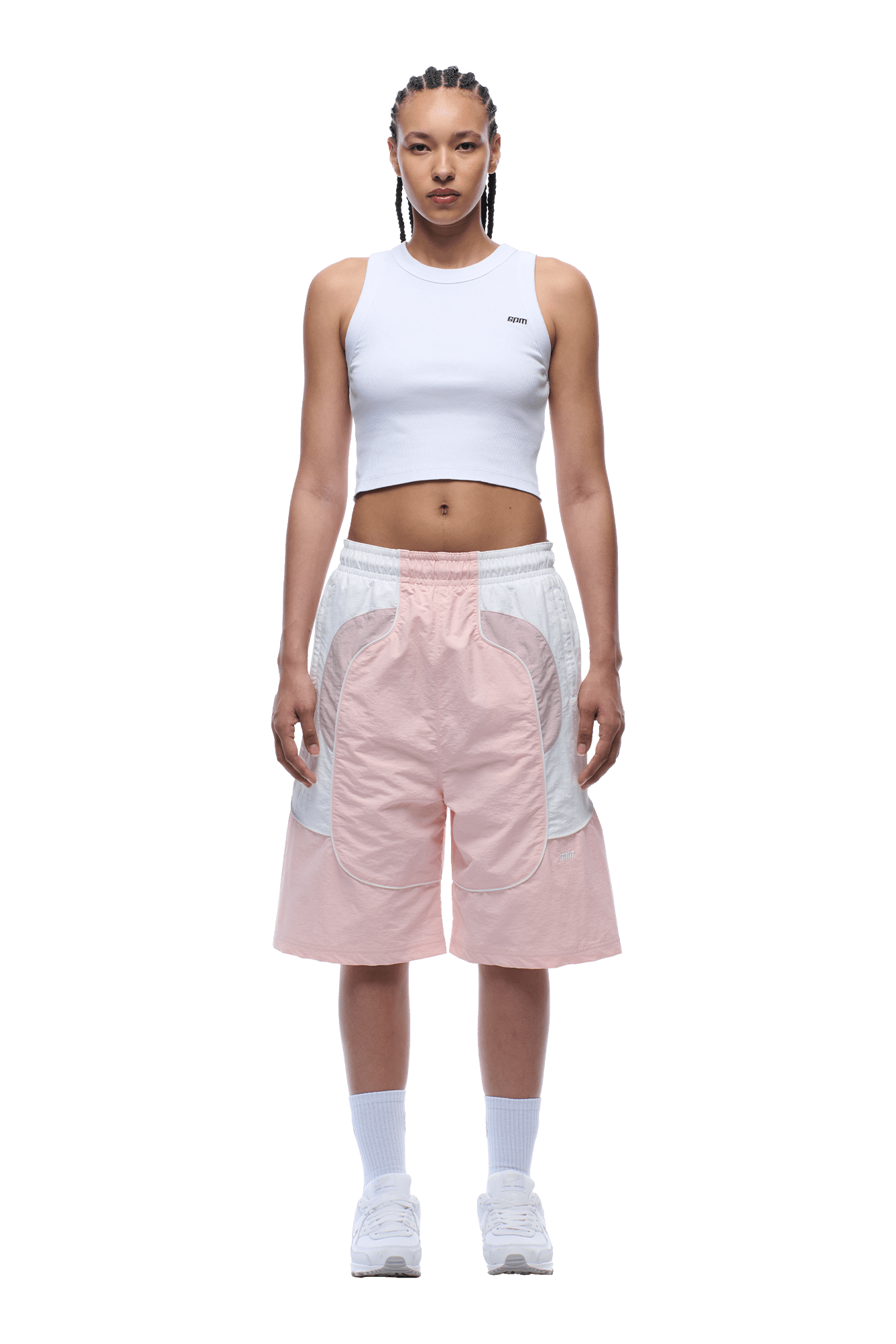 A person with braided hair stands facing forward against a green background, wearing a white sleeveless crop top, 6PM BAGGY WARM UP SHORTS PINK, white socks, and white sneakers.