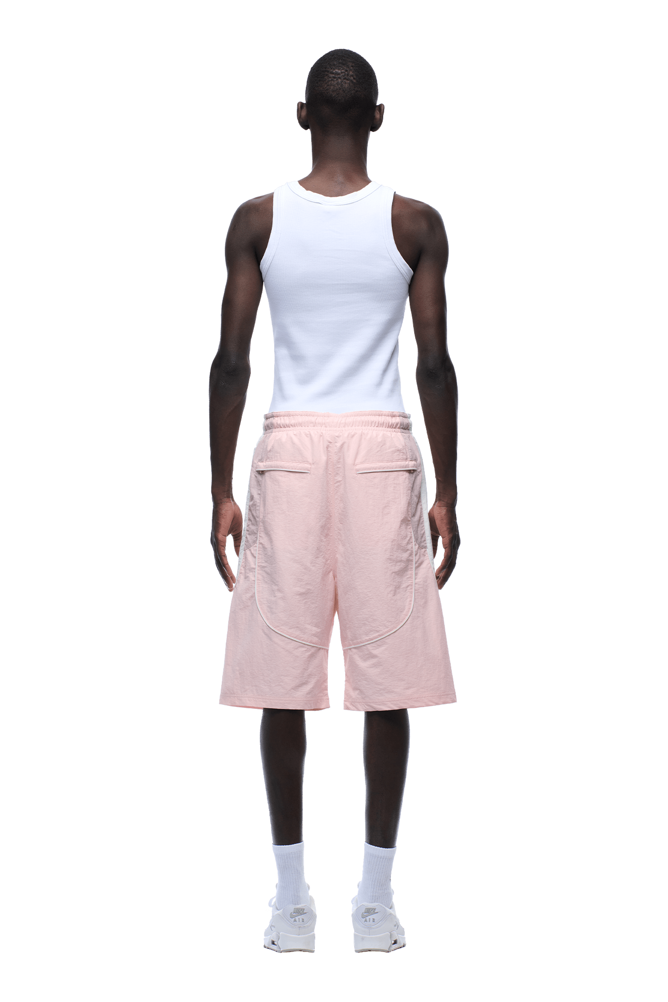 A person faces away, wearing 6PM BAGGY WARM UP SHORTS PINK with a white tank top, white socks, and white sneakers against a plain background.