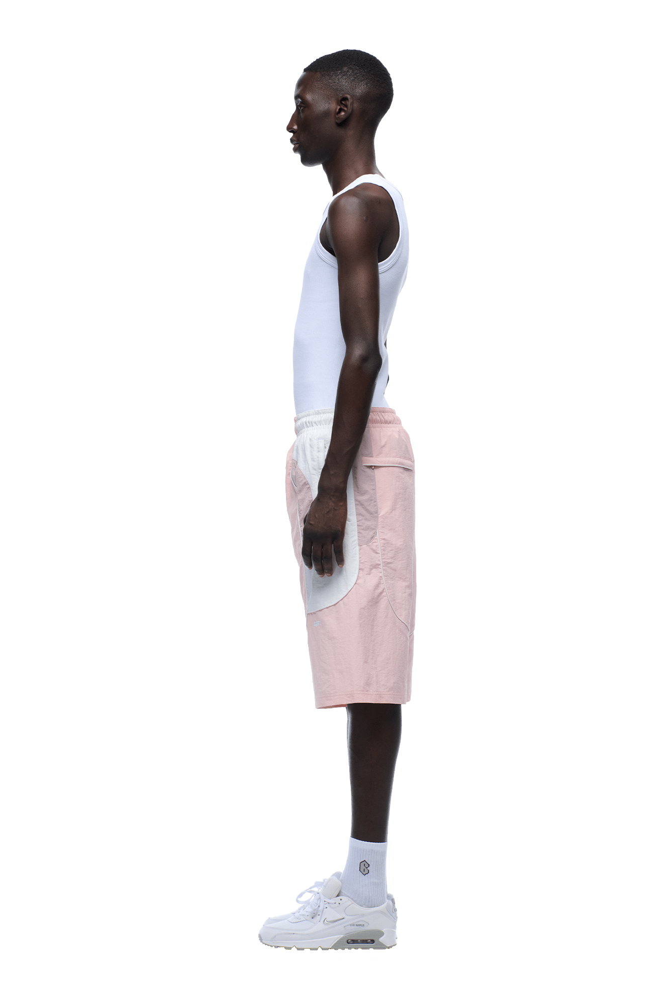 A man in profile wears a white tank top, 6PM BAGGY WARM UP SHORTS PINK with white pockets, white socks, and sneakers against a plain background.