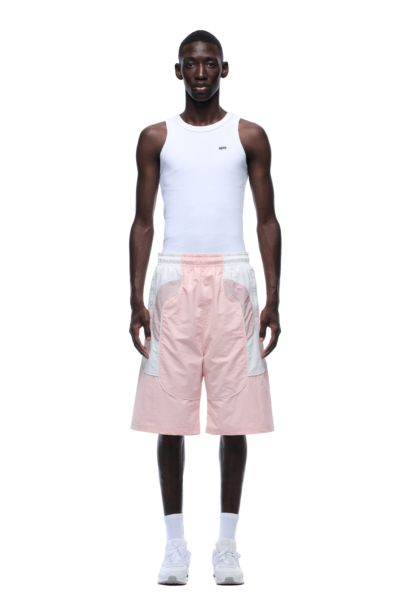 A man stands facing forward against a plain green background, wearing a fitted white tank top, 6PM BAGGY WARM UP SHORTS PINK, white socks, and white sneakers.