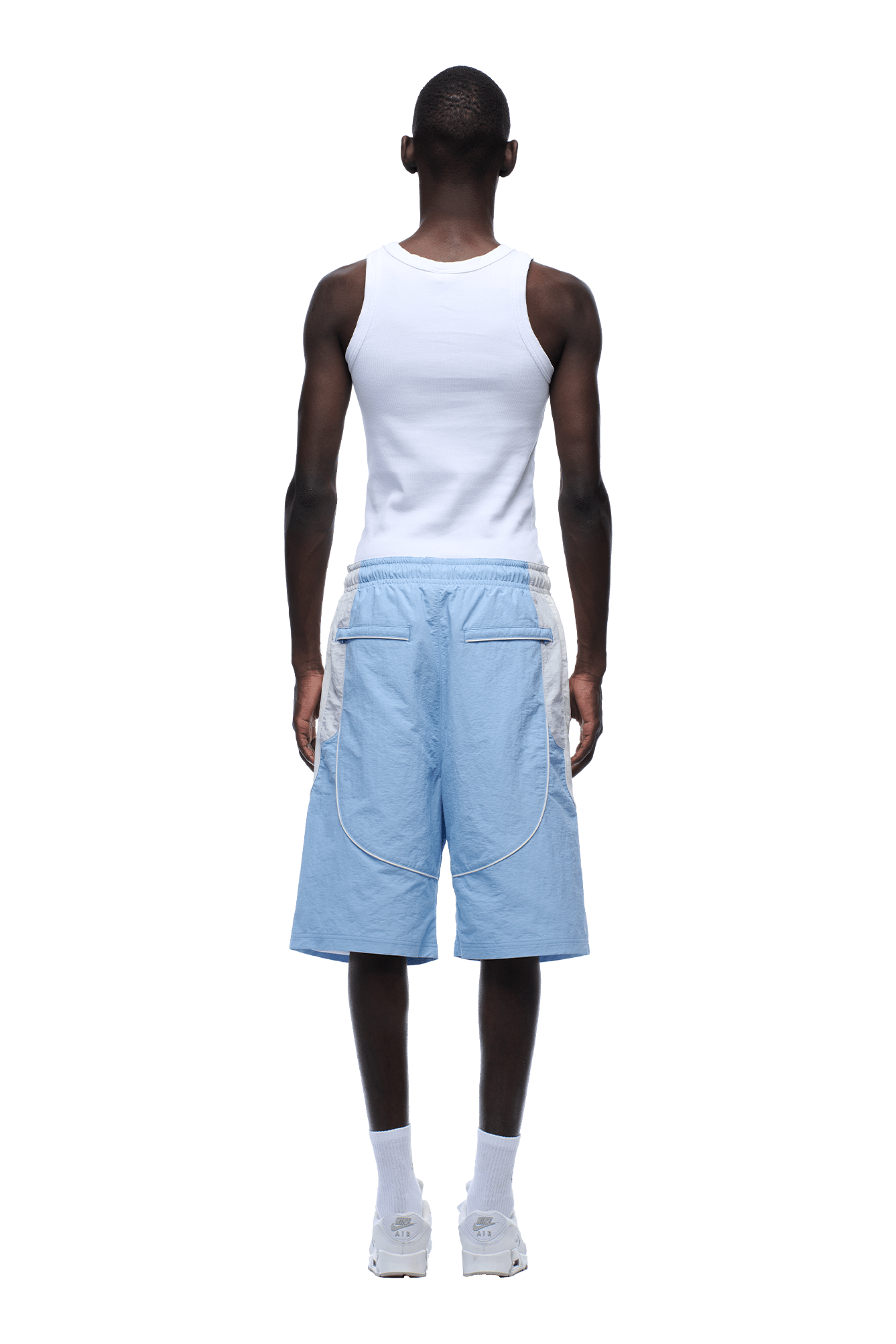 A person stands facing away on a plain green background, wearing a white tank top and 6PM BAGGY WARM UP SHORTS ICE BLUE, paired with white socks and sneakers.