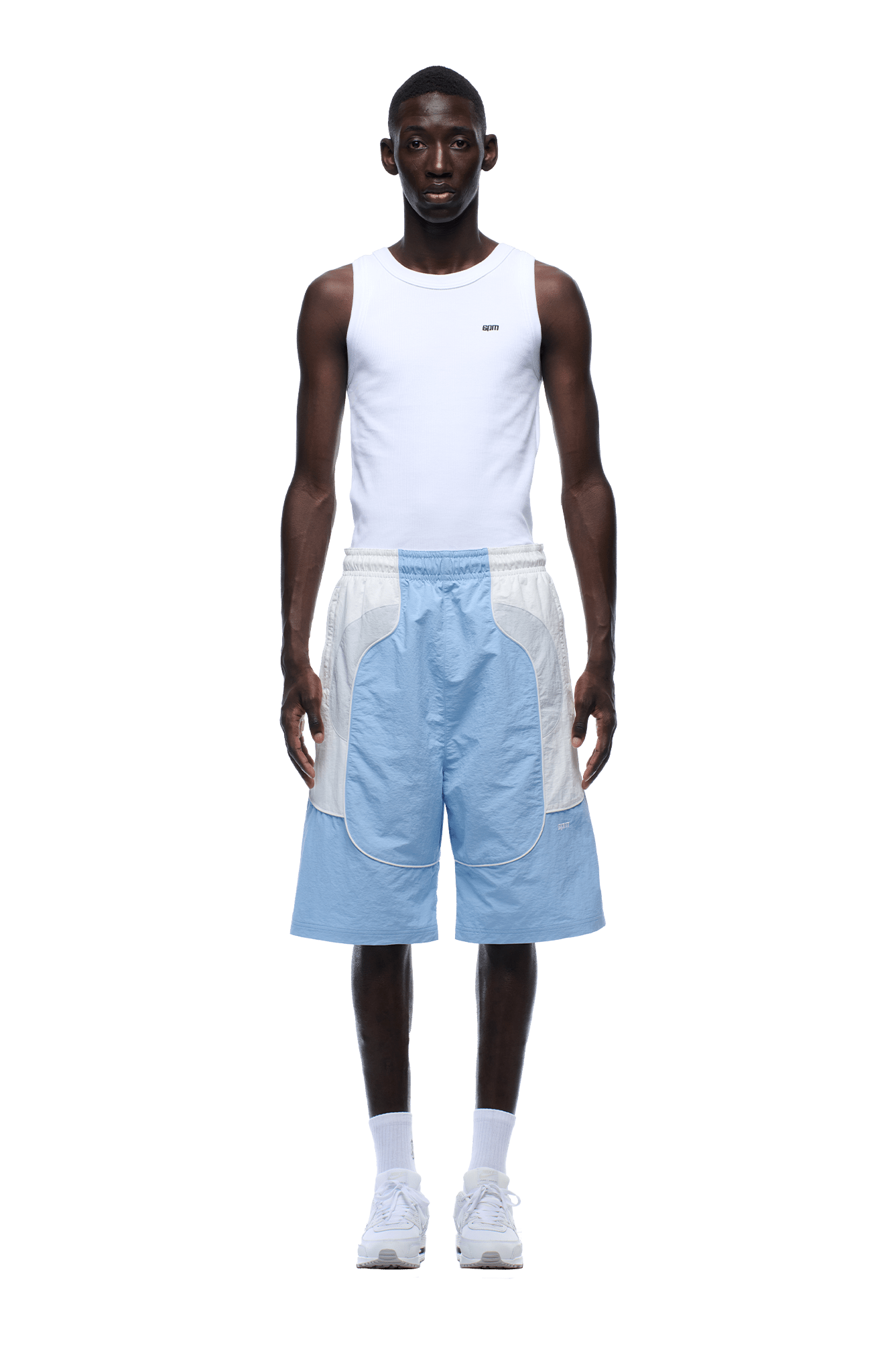A man stands facing forward in a white sleeveless tank top, 6PM BAGGY WARM UP SHORTS ICE BLUE, white socks, and white sneakers against a plain background.