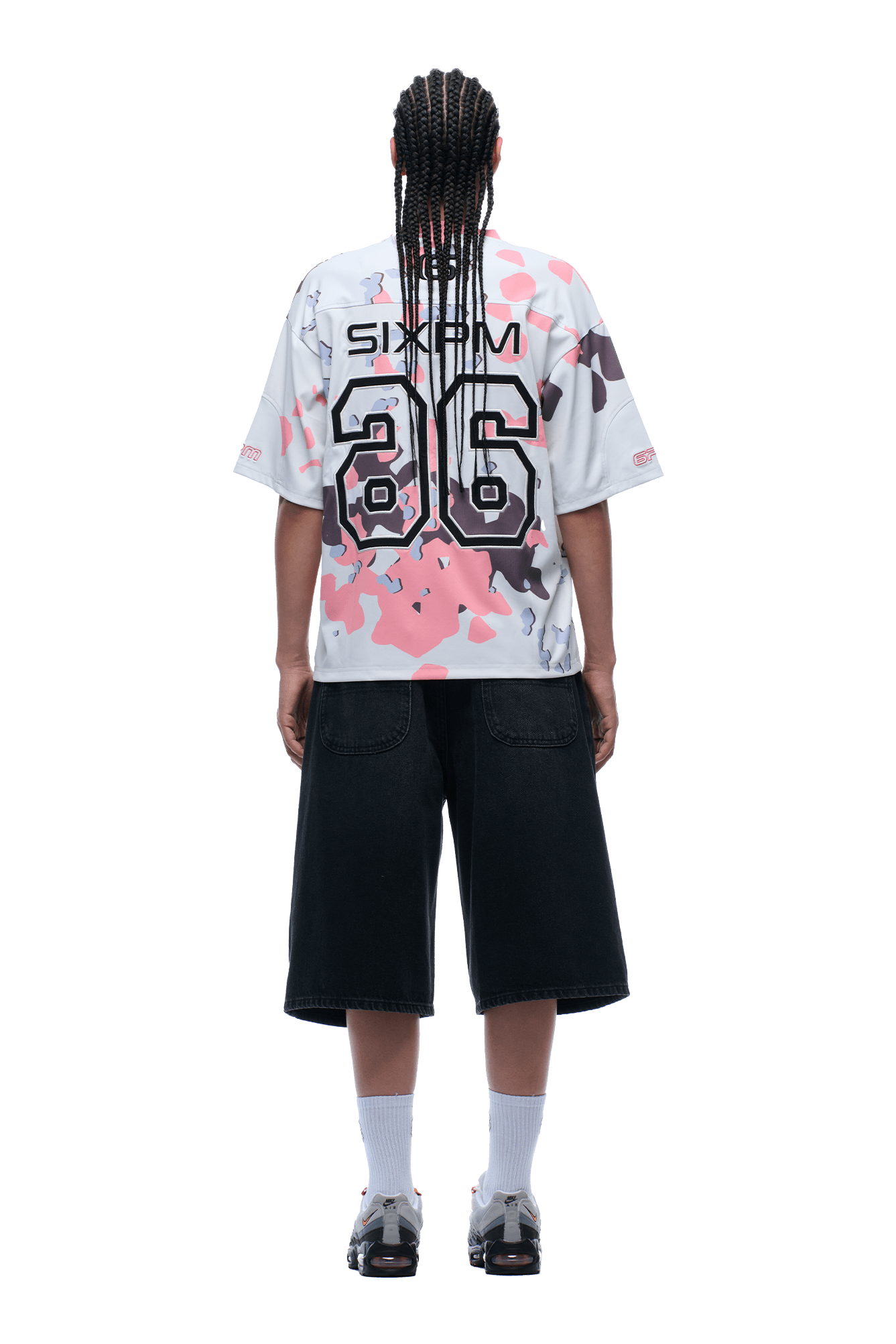 A person with braided hair, wearing the 6PM CAMO JERSEY FLAMINGO shirt, black knee-length shorts, white socks, and sneakers stands facing away from the camera.