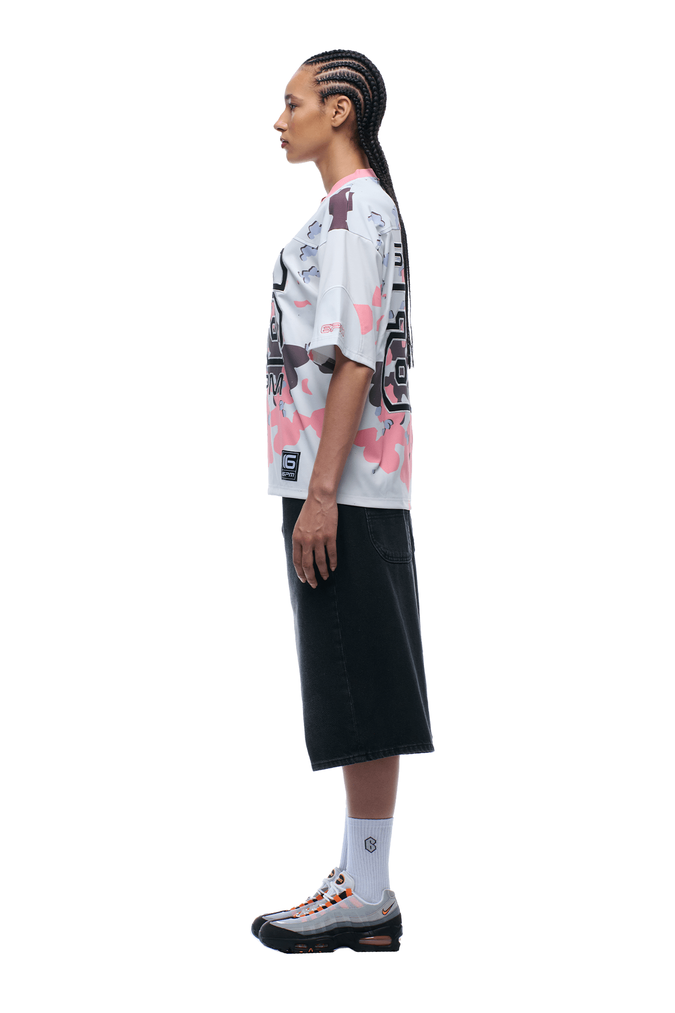 A woman with braided hair stands in profile against a plain background, wearing the 6PM CAMO JERSEY FLAMINGO patterned t-shirt, a black midi skirt, white socks, and sneakers.