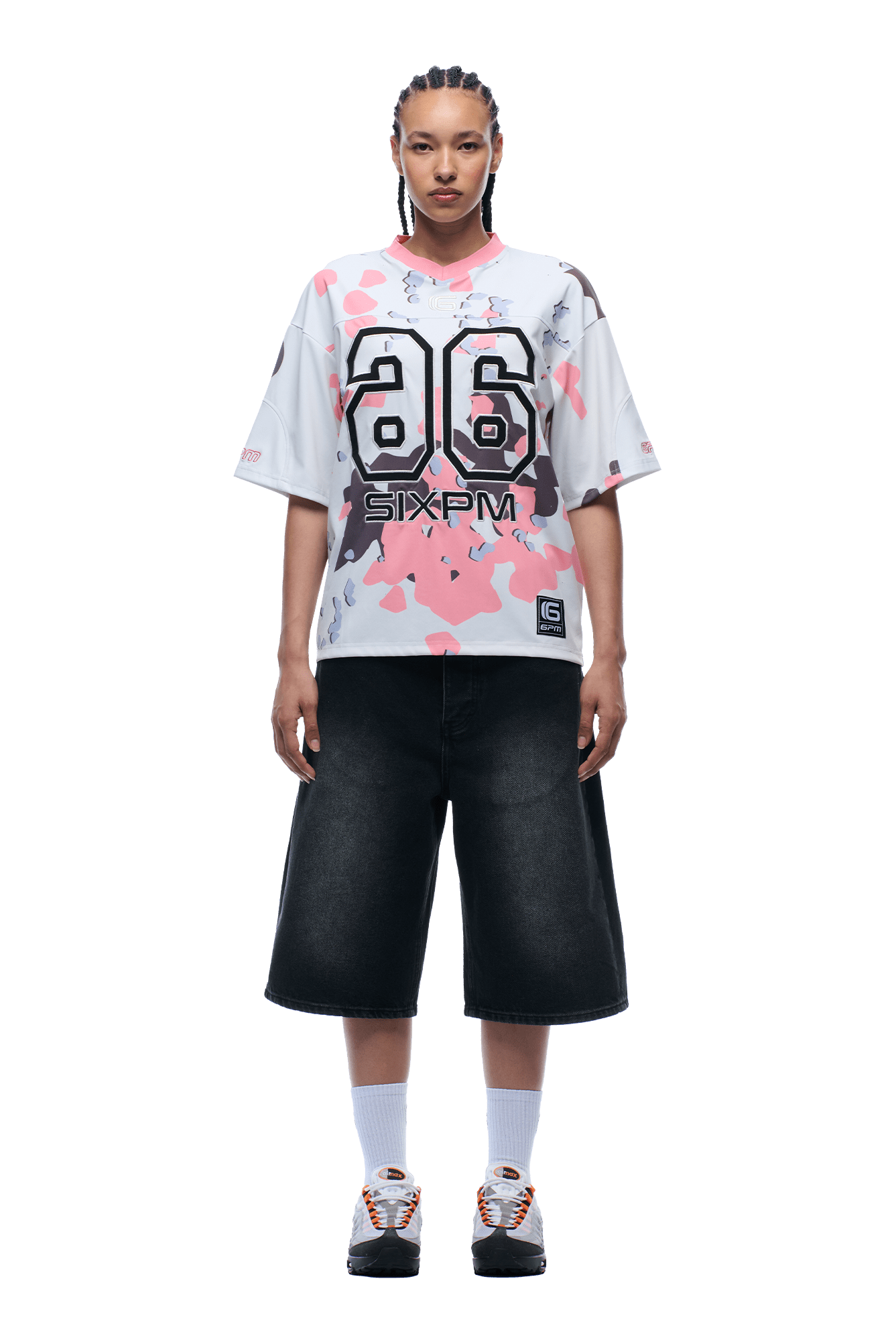 A person with braided hair wears the 6PM CAMO JERSEY FLAMINGO, wide black knee-length shorts, white socks, and sneakers, standing facing forward against a plain background.