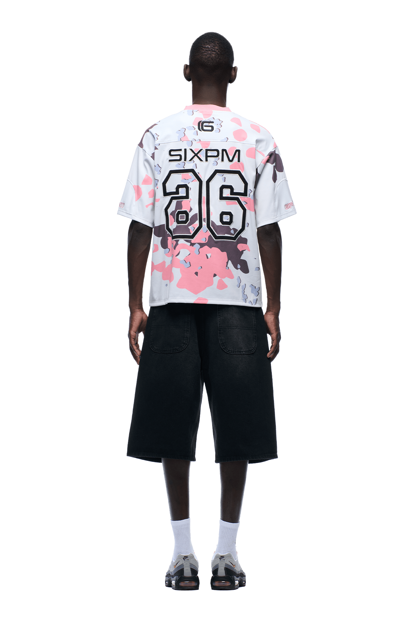 A person stands facing away, wearing the 6PM CAMO JERSEY FLAMINGO—a white shirt with pink, gray, and black splashes, SIXPM and 86 on the back—paired with black knee-length shorts, white socks, and gray sandals.