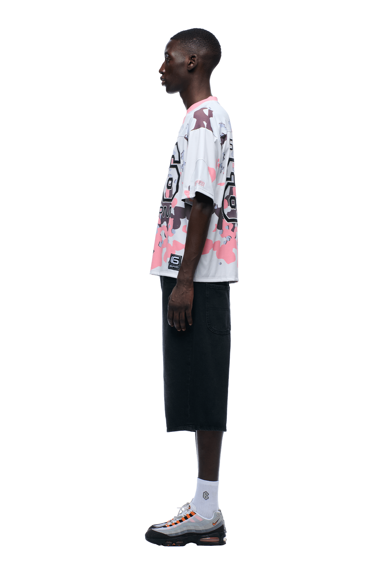 A person stands in profile against a plain background, wearing the 6PM CAMO JERSEY FLAMINGO—a white and pink patterned oversized T-shirt—paired with black knee-length shorts, white socks, and gray sneakers.