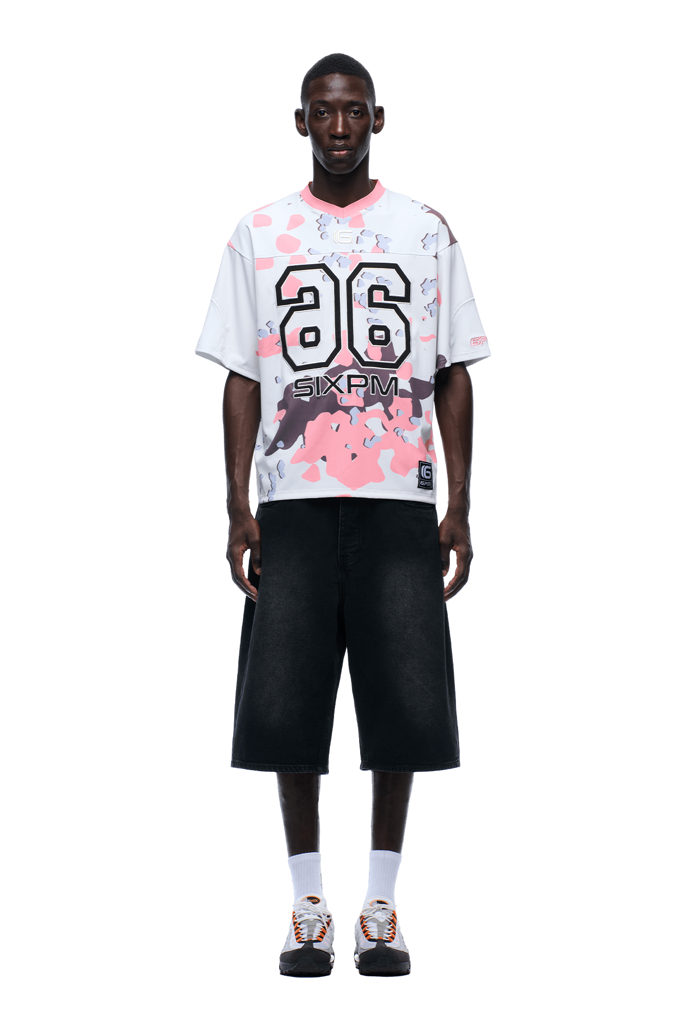 A man faces forward, wearing the 6PM CAMO JERSEY FLAMINGO—a white and pink graphic jersey with the number 26—paired with wide black knee-length shorts, white socks, and white sneakers with orange accents.