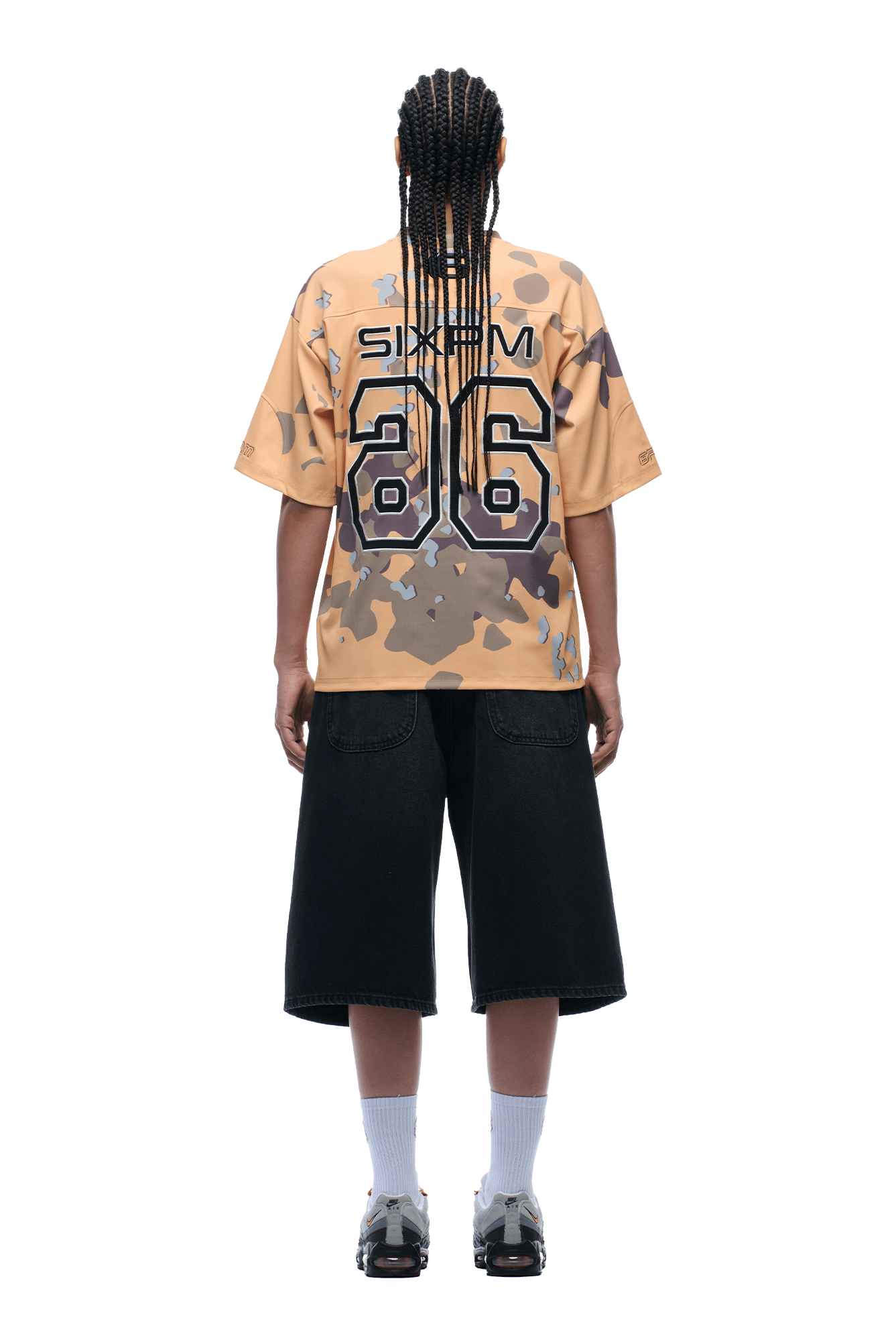 A person with long braids stands facing away, wearing the 6PM CAMO JERSEY BURRO with 86 on the back, black knee-length shorts, white socks, and gray sneakers, against a transparent background.
