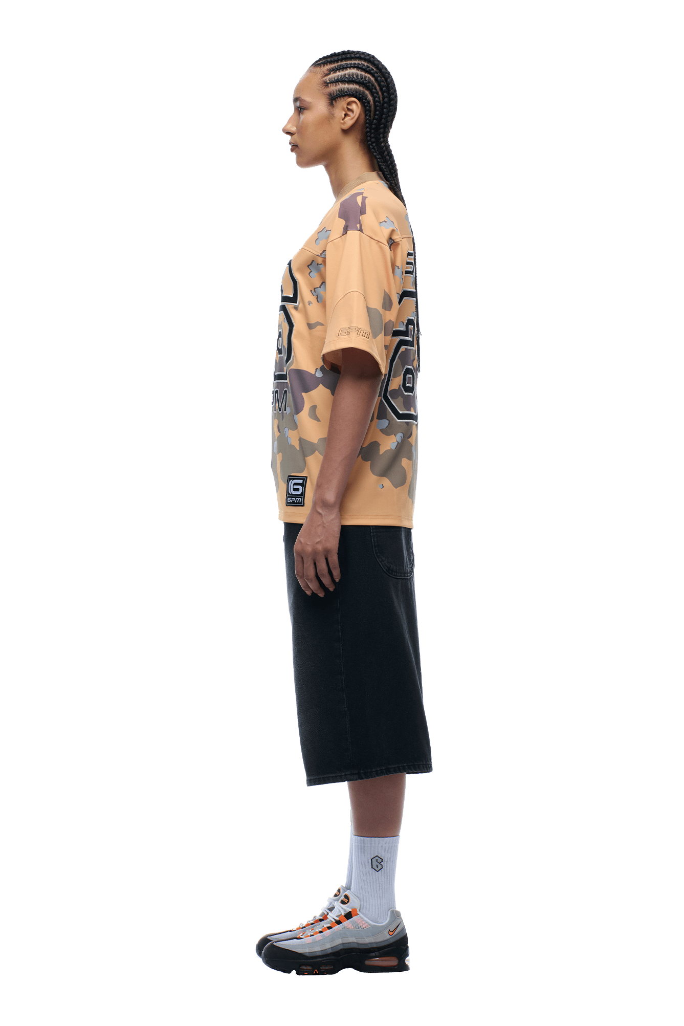 Against a plain backdrop, a person with braided hair stands in profile wearing the 6PM CAMO JERSEY BURRO shirt, paired with a black knee-length skirt, white socks, and sneakers.