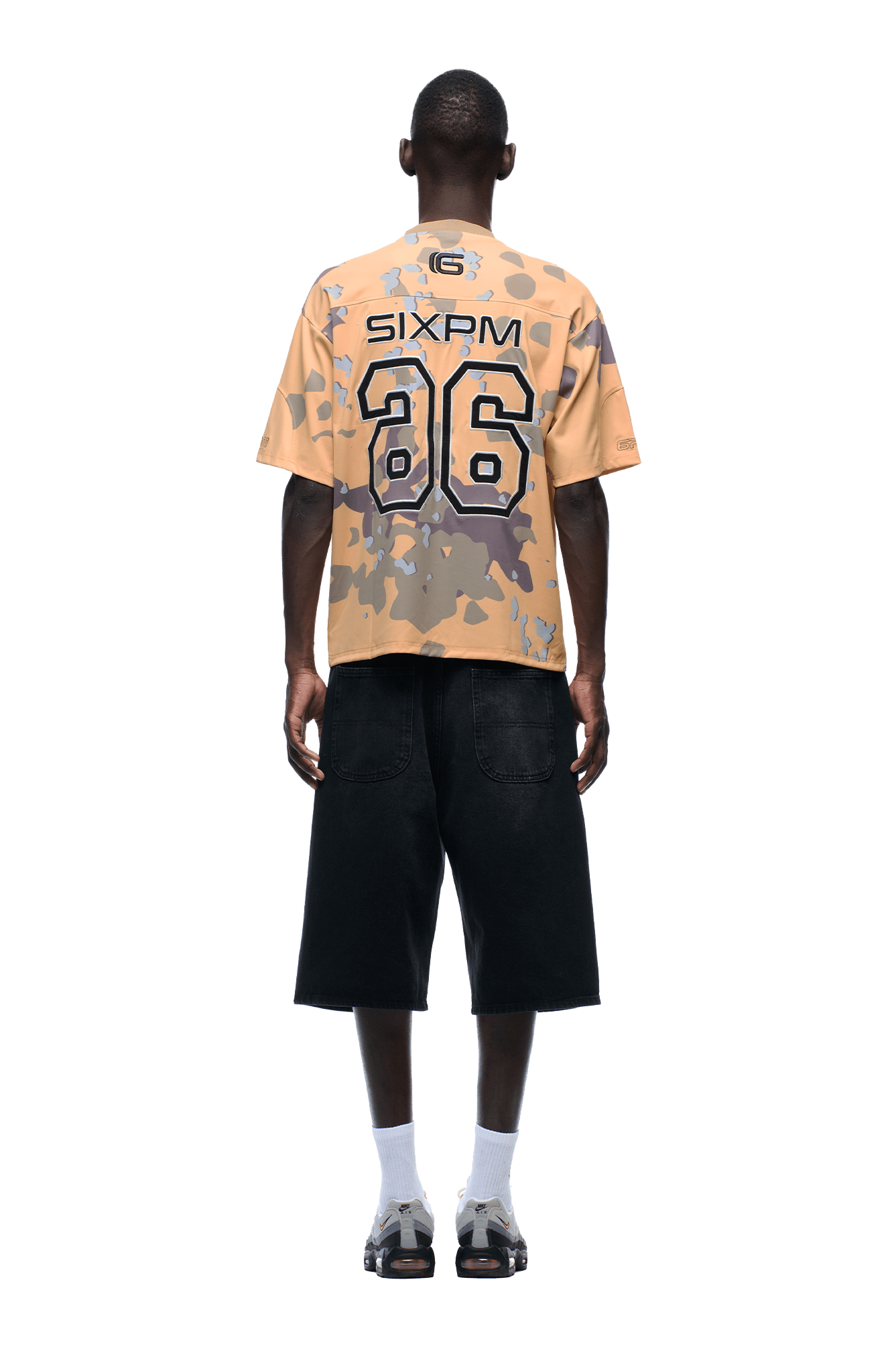 Against a plain background, a person wears the CAMO JERSEY BURRO by 6PM with 86 on the back, loose black knee-length shorts, white socks, and beige sneakers, standing facing away.