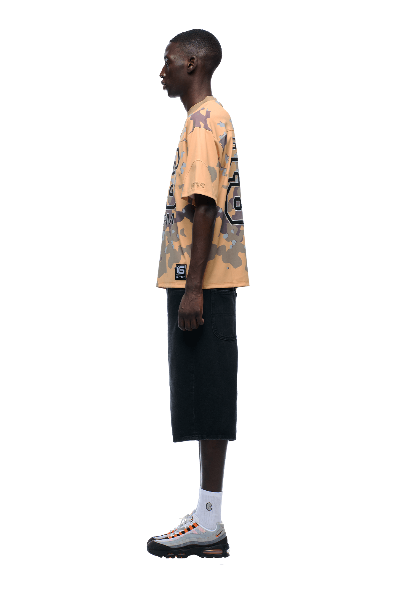 A person stands in profile against a plain background, wearing the 6PM CAMO JERSEY BURRO t-shirt, black knee-length shorts, white socks, and white sneakers with orange and black accents.
