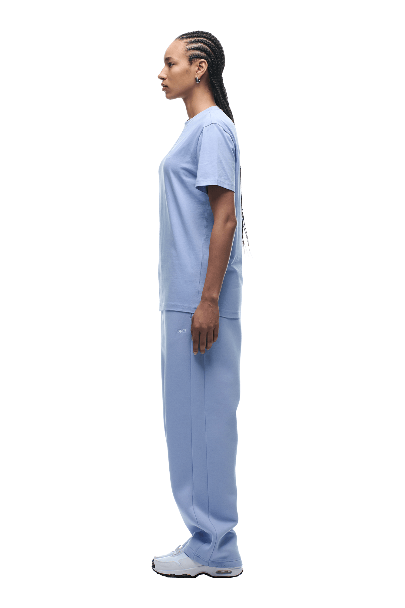 A woman with long braids stands in profile, showcasing the 6PM T-SHIRT REGULAR FIT SOFT VIOLET with matching jogger pants and white sneakers against a plain background.