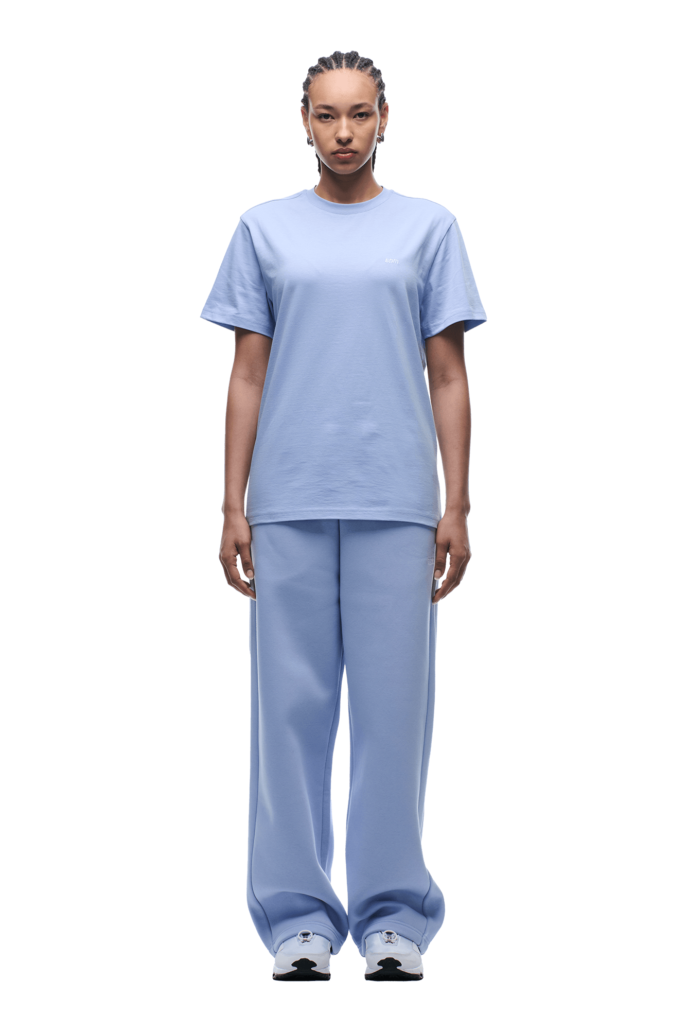 A person with neat braids and a neutral expression faces forward, wearing the 6PM T-SHIRT REGULAR FIT SOFT VIOLET and matching loose pants with white sneakers. The background is transparent for a clean, stylish look.