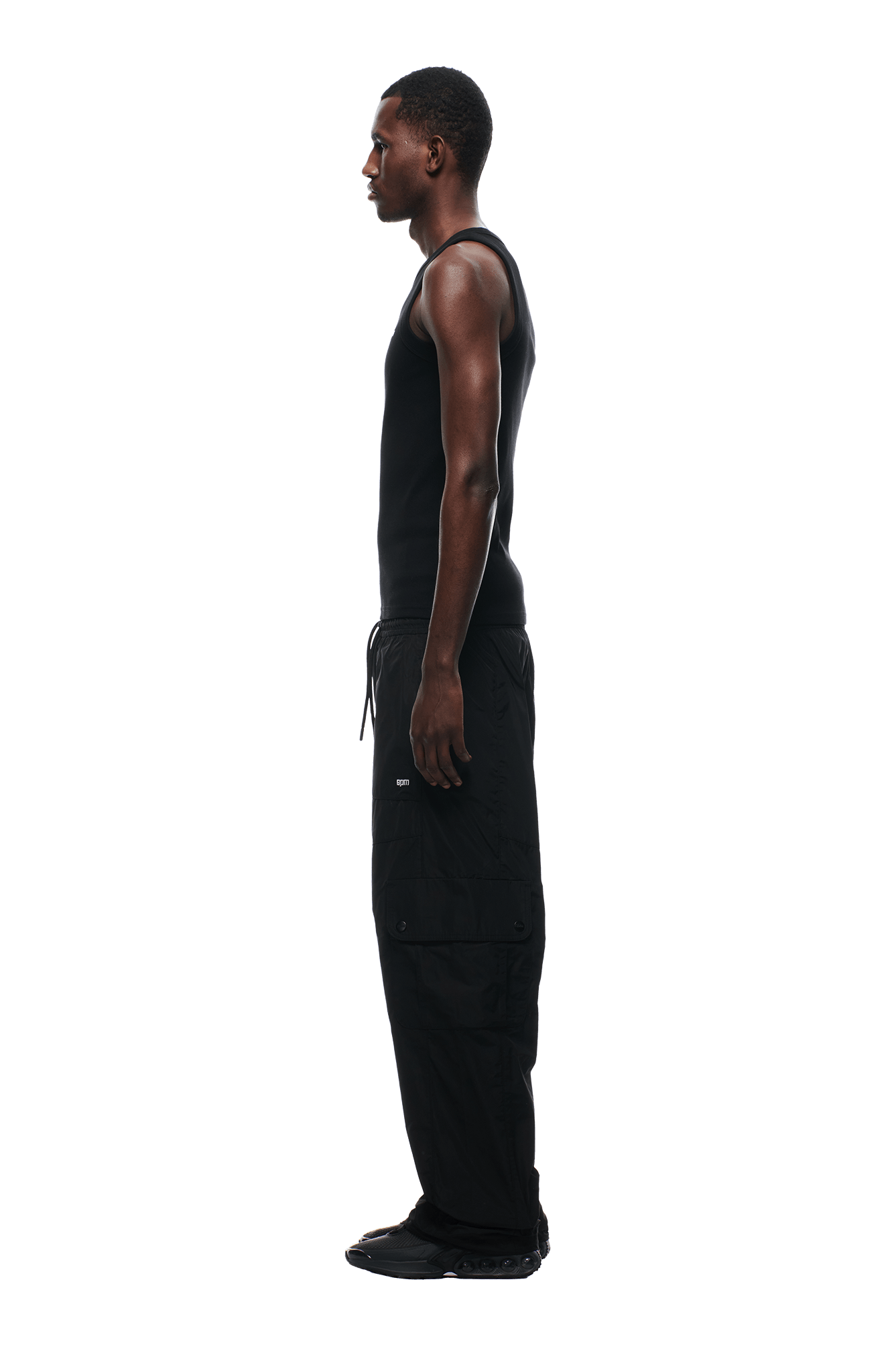 A man stands in side profile, wearing the 6PM NYLON CARGO PANTS BLACK with a black tank top and black shoes, against a plain background.