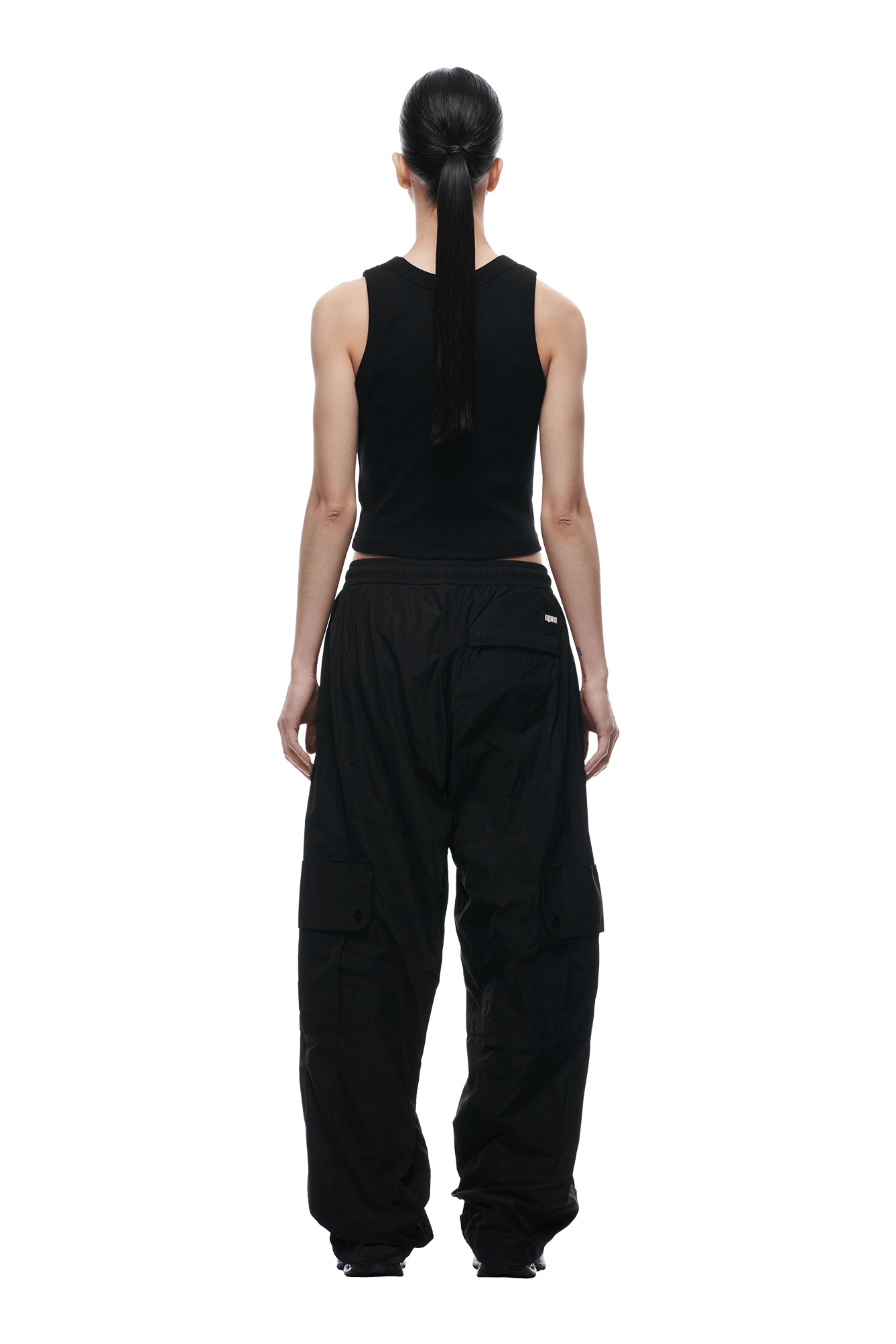 Someone with long dark hair in a ponytail faces away from the camera, wearing 6PM NYLON CARGO PANTS BLACK and a black sleeveless top, set against a green background.