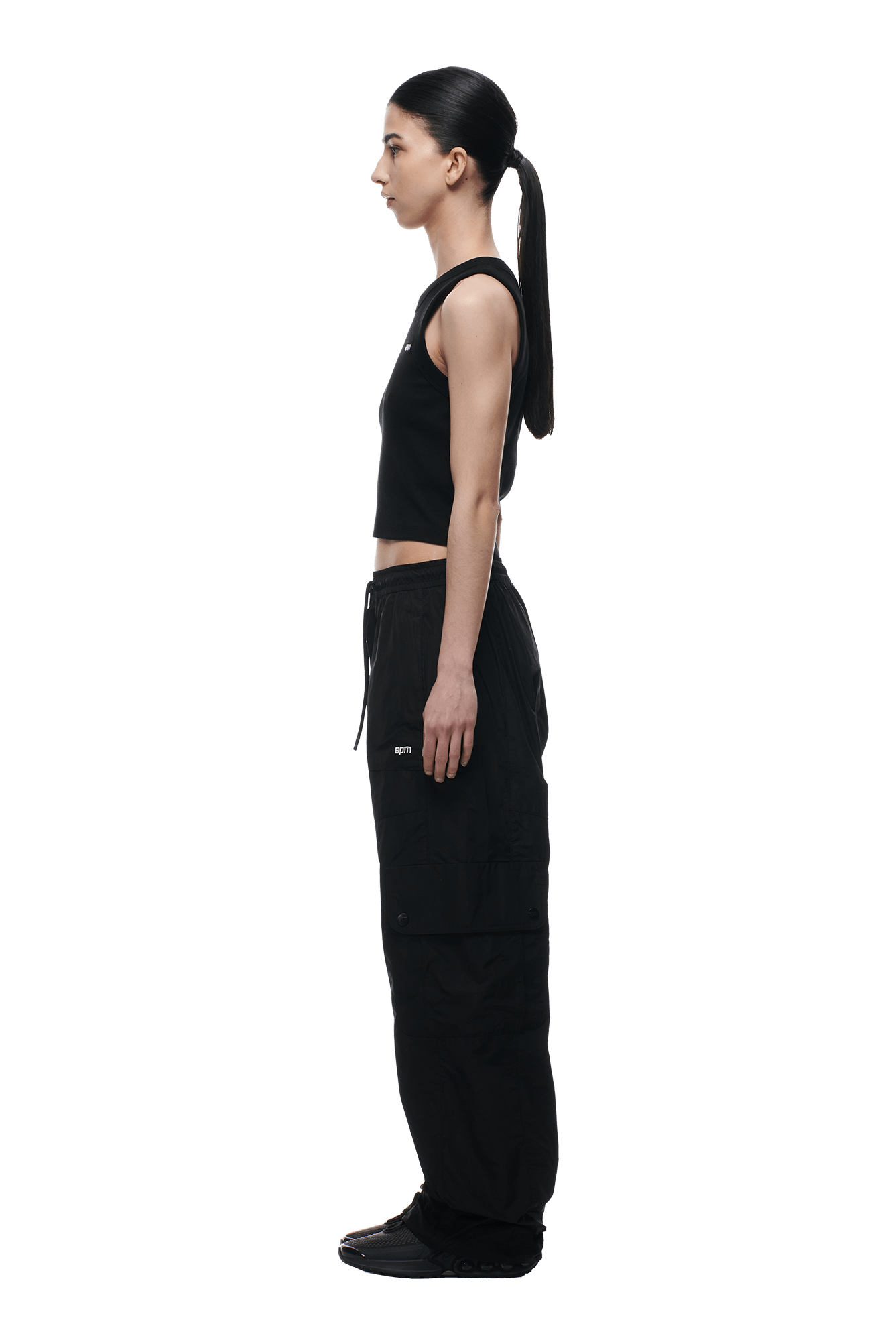A woman with long dark hair in a ponytail stands in profile against a transparent background, wearing 6PM NYLON CARGO PANTS BLACK, a sleeveless black top, and black shoes.