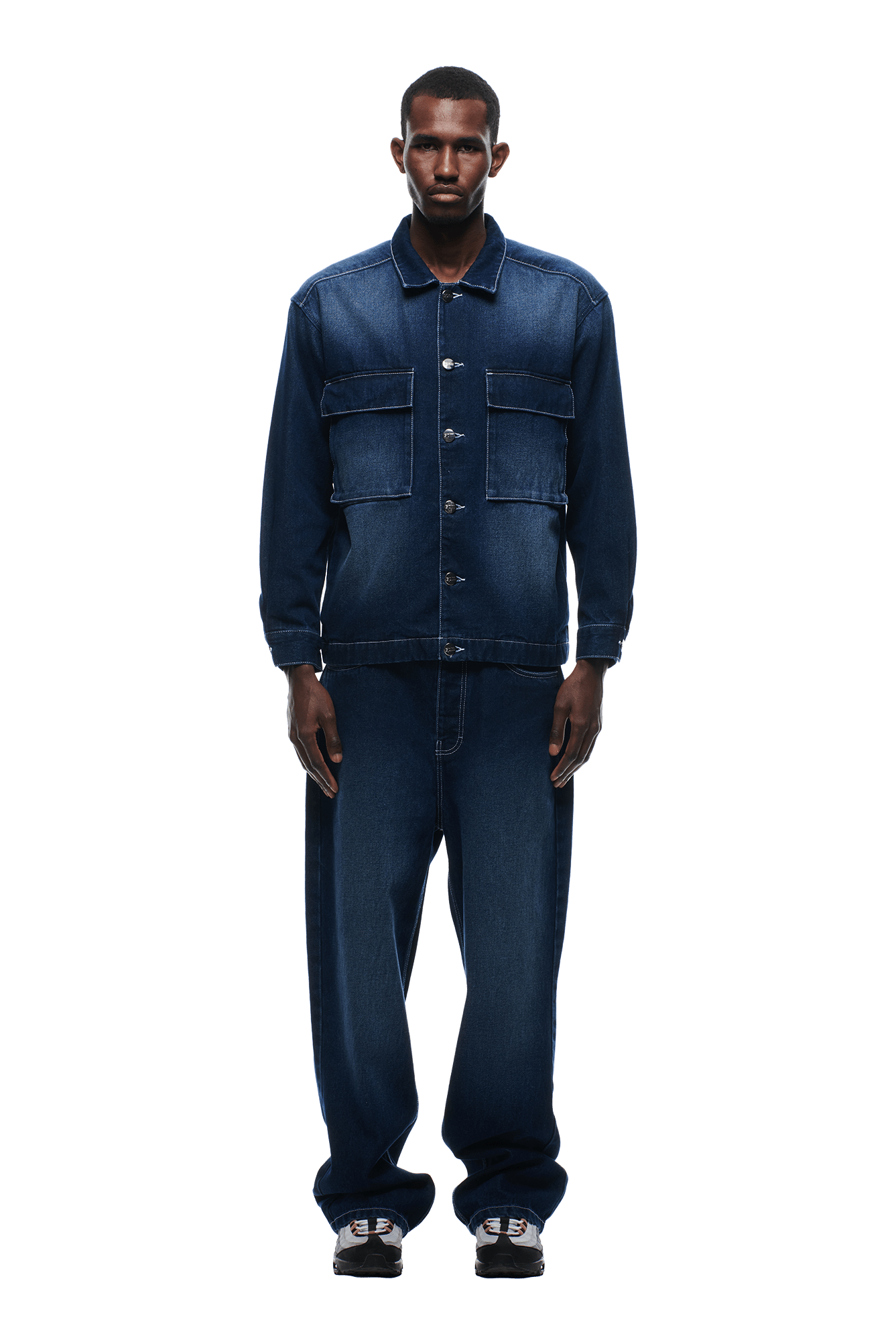 A man faces forward in a 6PM SCRIPT LOGO DENIM JACKET DARK BLUE with large front pockets and matching loose-fit jeans, paired with black and white sneakers, against a plain white background.