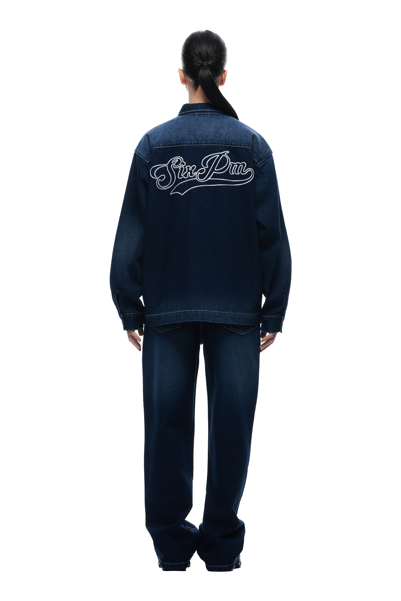 A person with dark hair in a ponytail wears the 6PM SCRIPT LOGO DENIM JACKET DARK BLUE with matching jeans. The jacket’s back displays large cursive "Sine Pru" embroidery. The background is plain white.