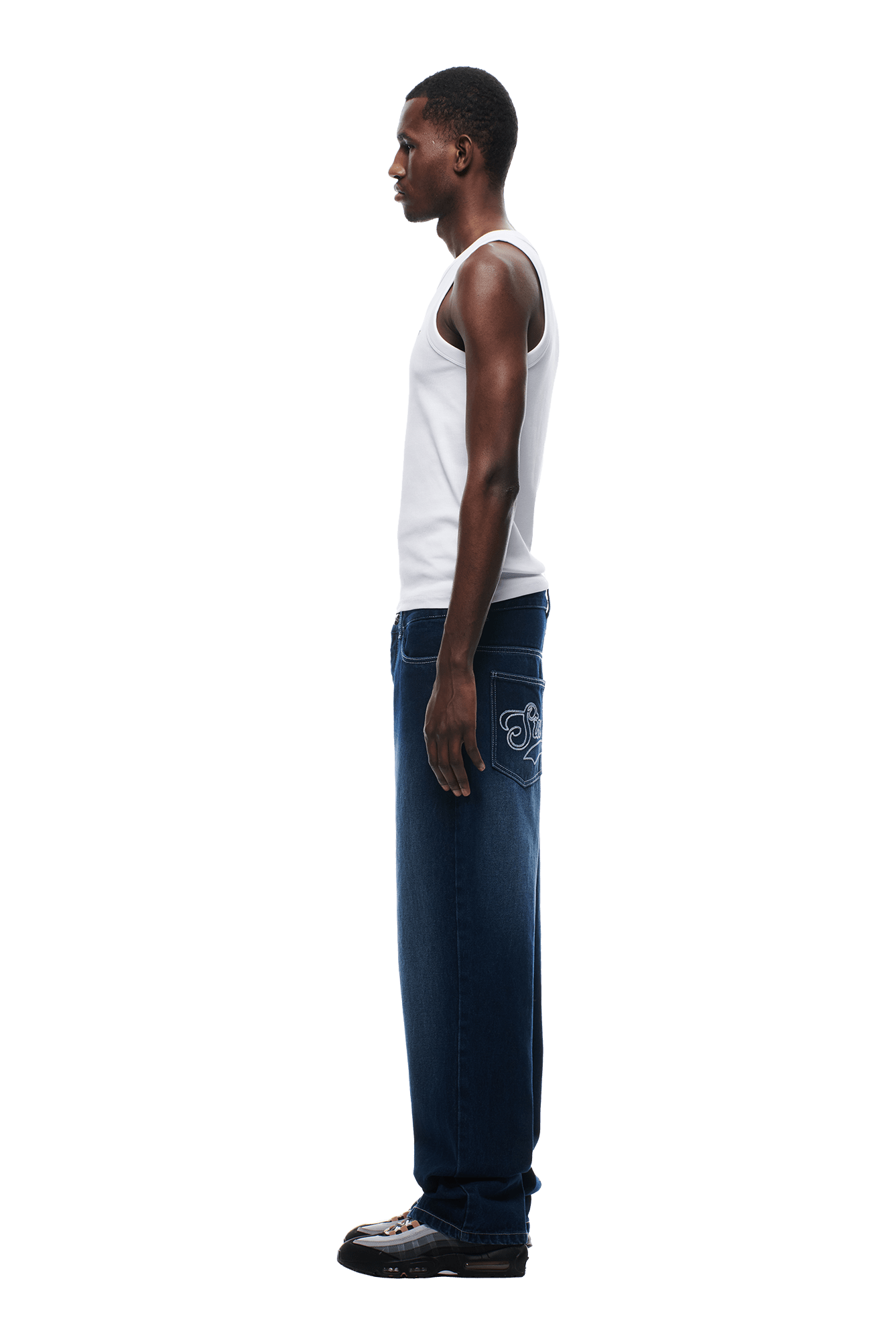 A man stands in profile against a white background, wearing a white tank top, black shoes, and 6PM SCRIPT LOGO DENIM WASHED DARK BLUE jeans featuring an embroidered back pocket.